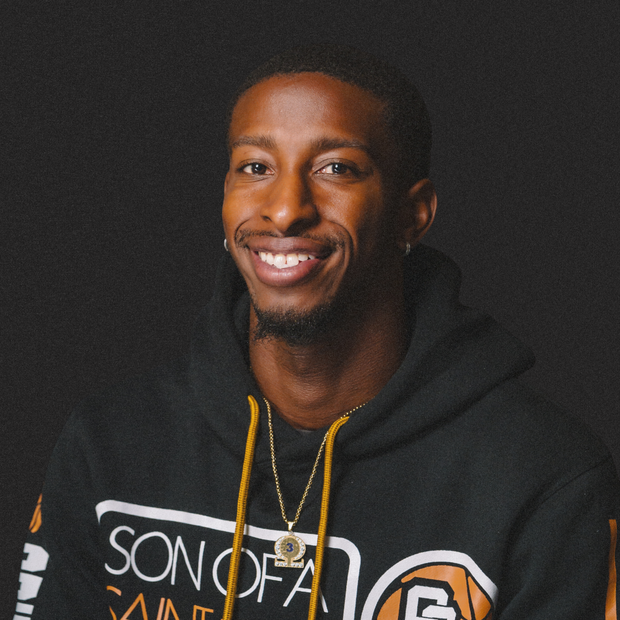 Portrait of a smiling young man with dark skin, short curly hair, wearing earrings, a black hoodie with orange and white text, and a gold necklace with a pendant, against a black background.
