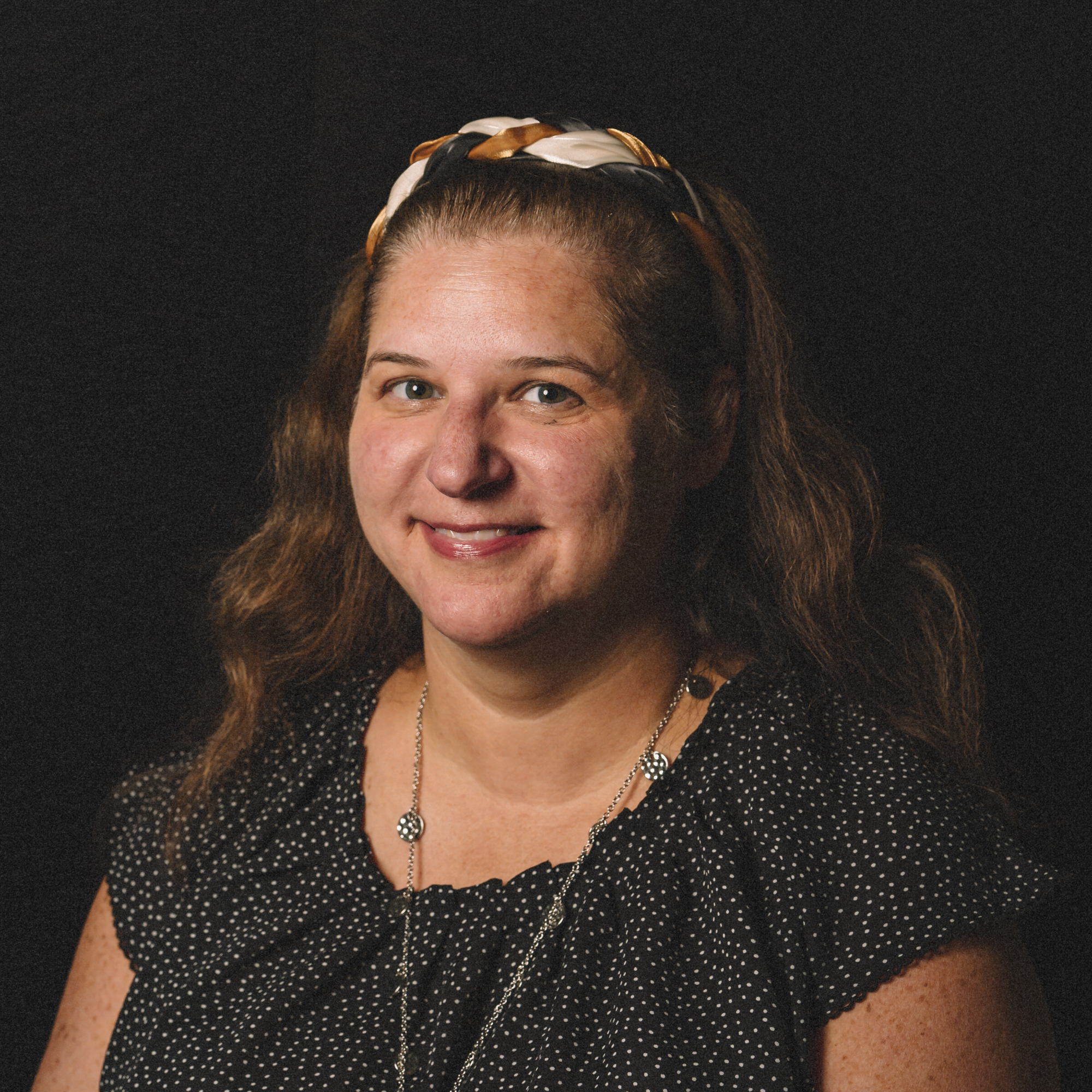 A woman with long wavy brown hair, wearing a black polka dot top and silver jewelry, smiling against a black background.