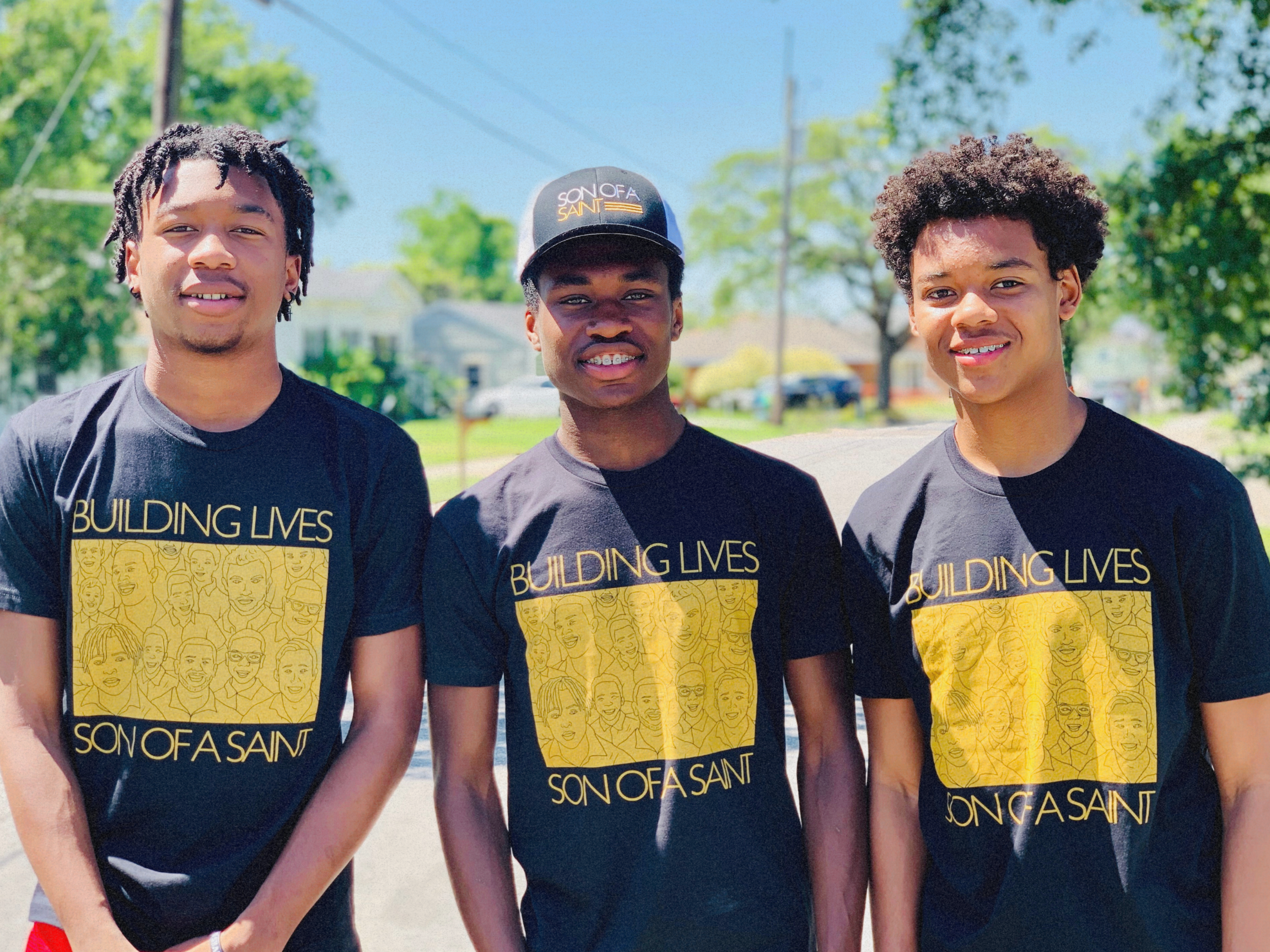 Three young men standing outdoors on a sunny day, wearing matching black T-shirts with yellow artwork and text that says, 'Building Lives Son of a Saint.' They are smiling and appear to be in a neighborhood with trees, houses, and parked cars in the background.