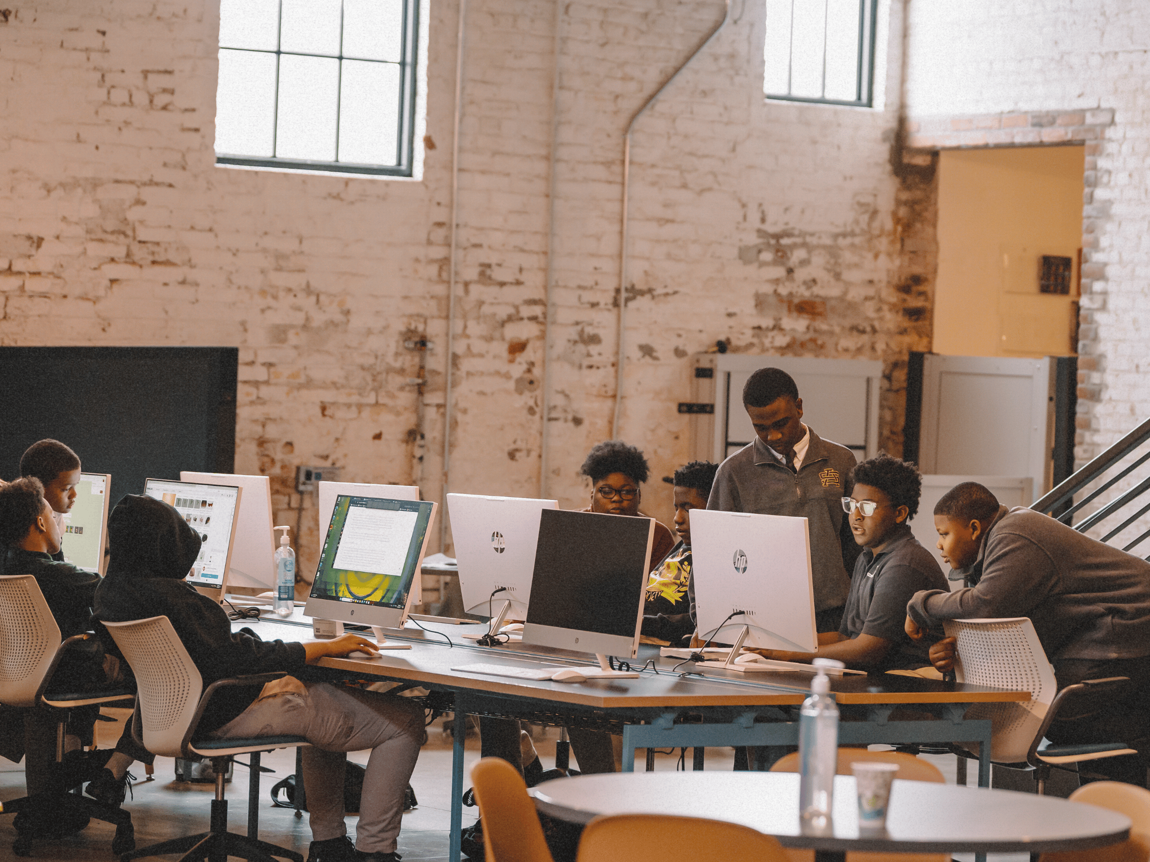Group of young students working together on computers in a modern classroom or workshop with exposed brick walls.