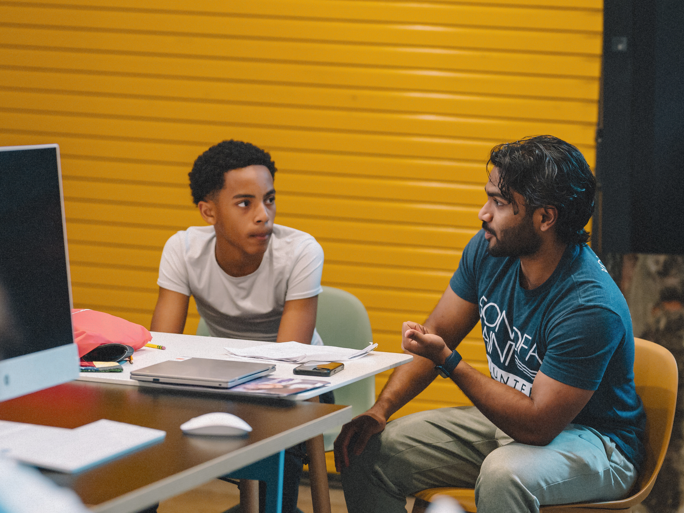 A young boy and an adult man sitting at a table having a conversation. The boy is looking at the man with a serious expression. The table has papers, a laptop, and an external mouse. The background features yellow horizontal wooden panels.
