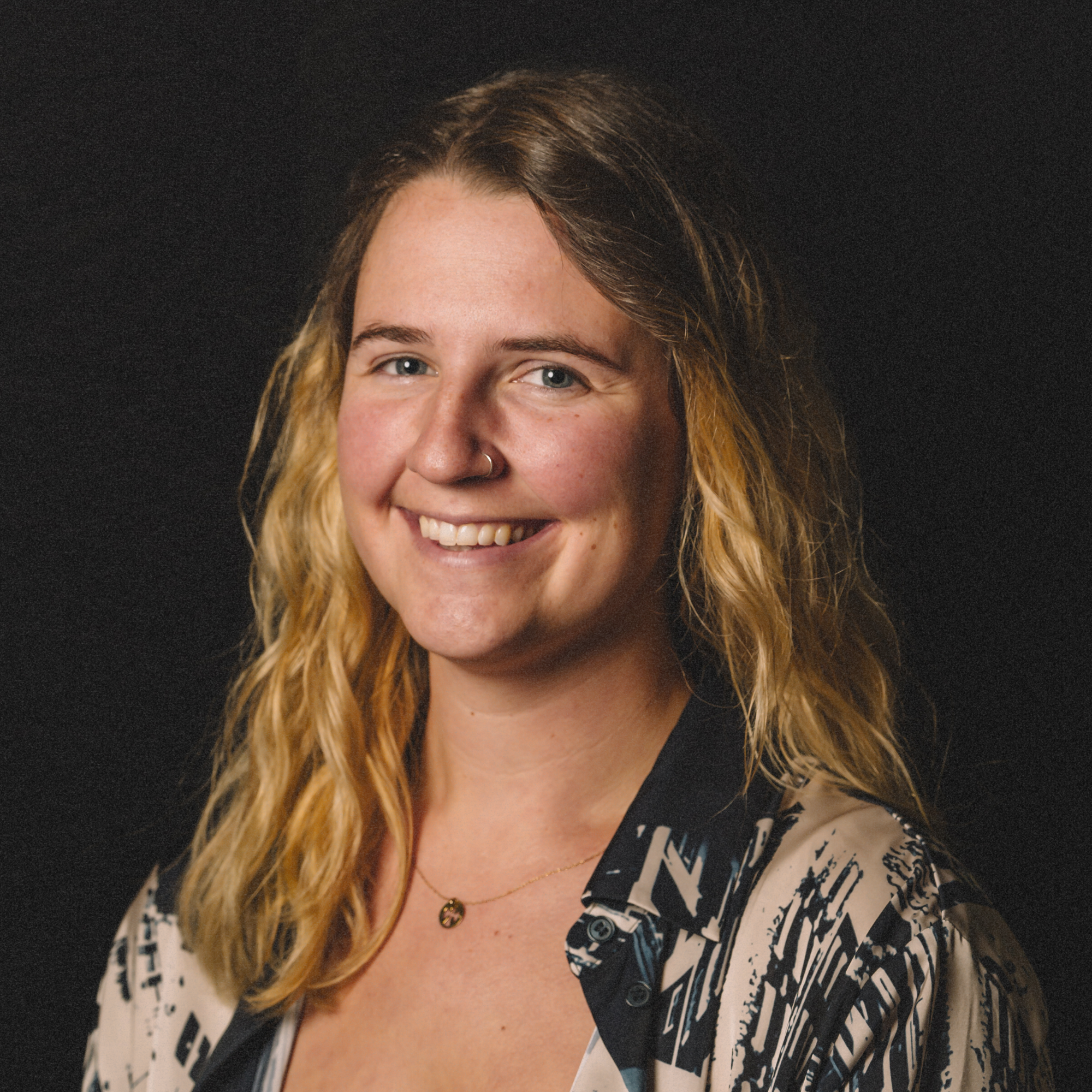 A smiling woman with blonde wavy hair wearing a black and white patterned shirt and a delicate necklace against a dark background.