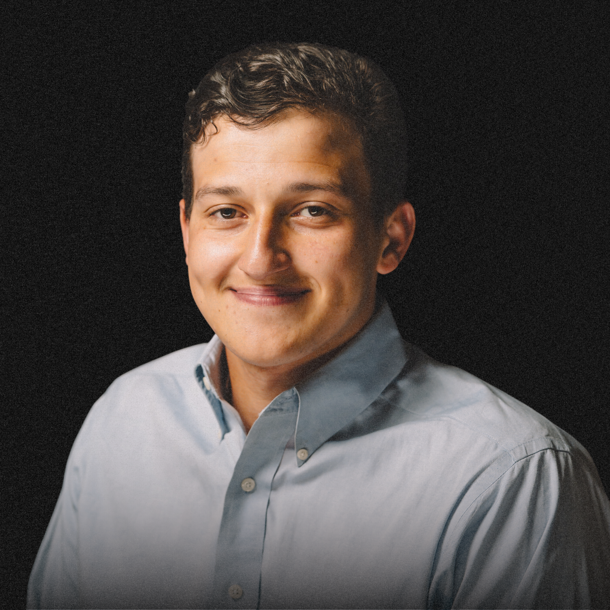 Portrait of a young man with dark hair and light skin, smiling, wearing a light gray collared shirt, against a black background.