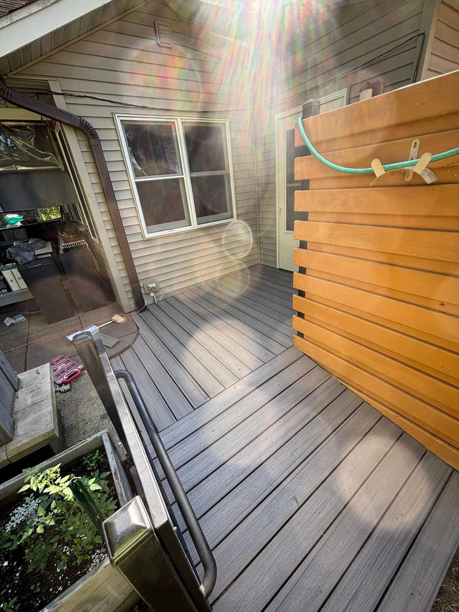 Sunlight shining onto a backyard wooden deck with a planter box containing green plants, a wooden privacy screen, a house wall with a window, a door, and a corner of a gas grill.