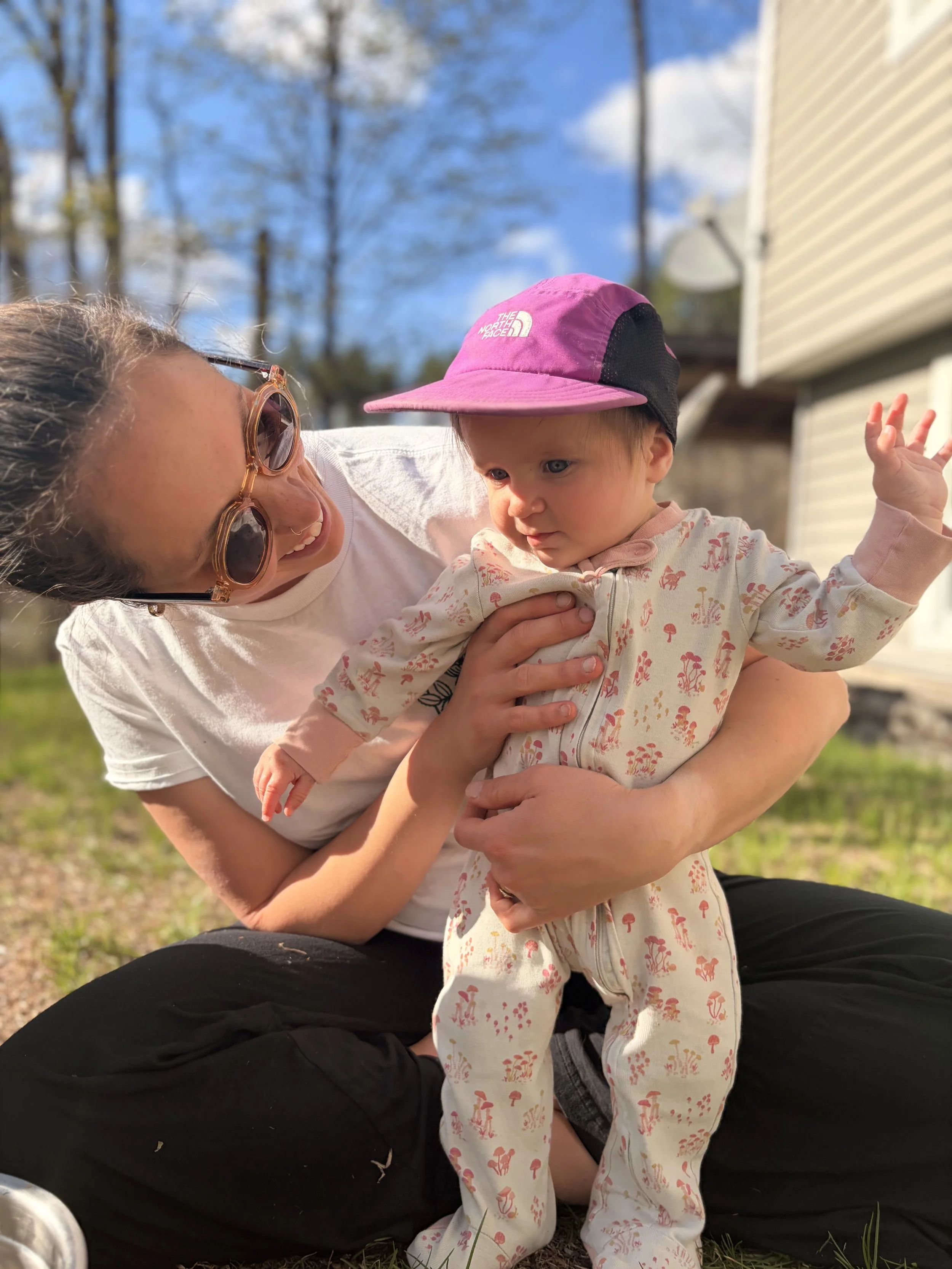 A woman with sunglasses and a white shirt is holding a young child outside on a sunny day. The child is wearing a pink and black hat and a cream-colored outfit with pink and yellow mushroom patterns. The woman is smiling and looking at the child, who is looking to the side with one hand raised. In the background, there are trees, a house, and a blue sky with some clouds.