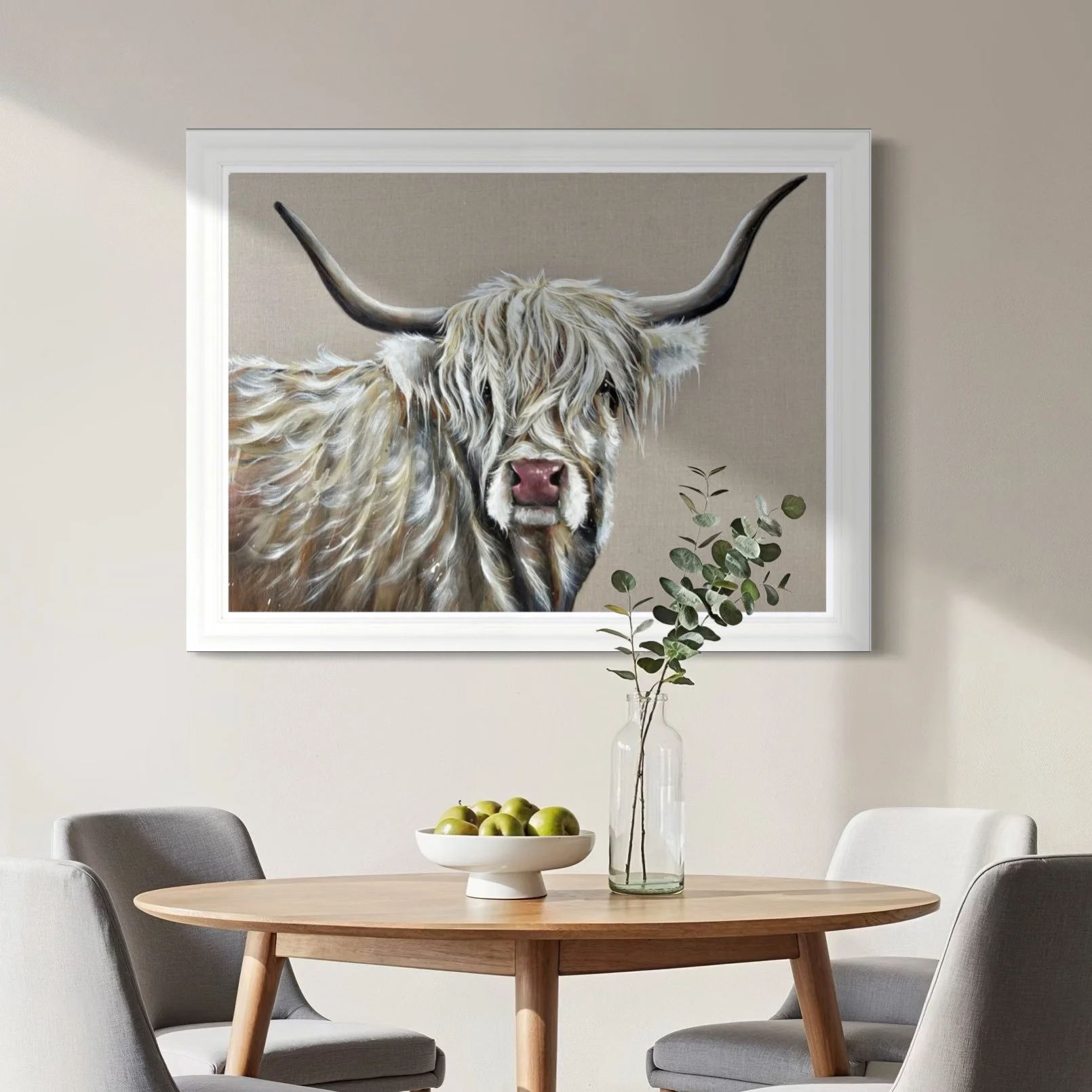 Framed highland cow print by Louise Luton. A dining room with a round wooden table surrounded by four upholstered chairs. On the table is a white bowl filled with green apples, and a tall glass vase with a leafy green branch.