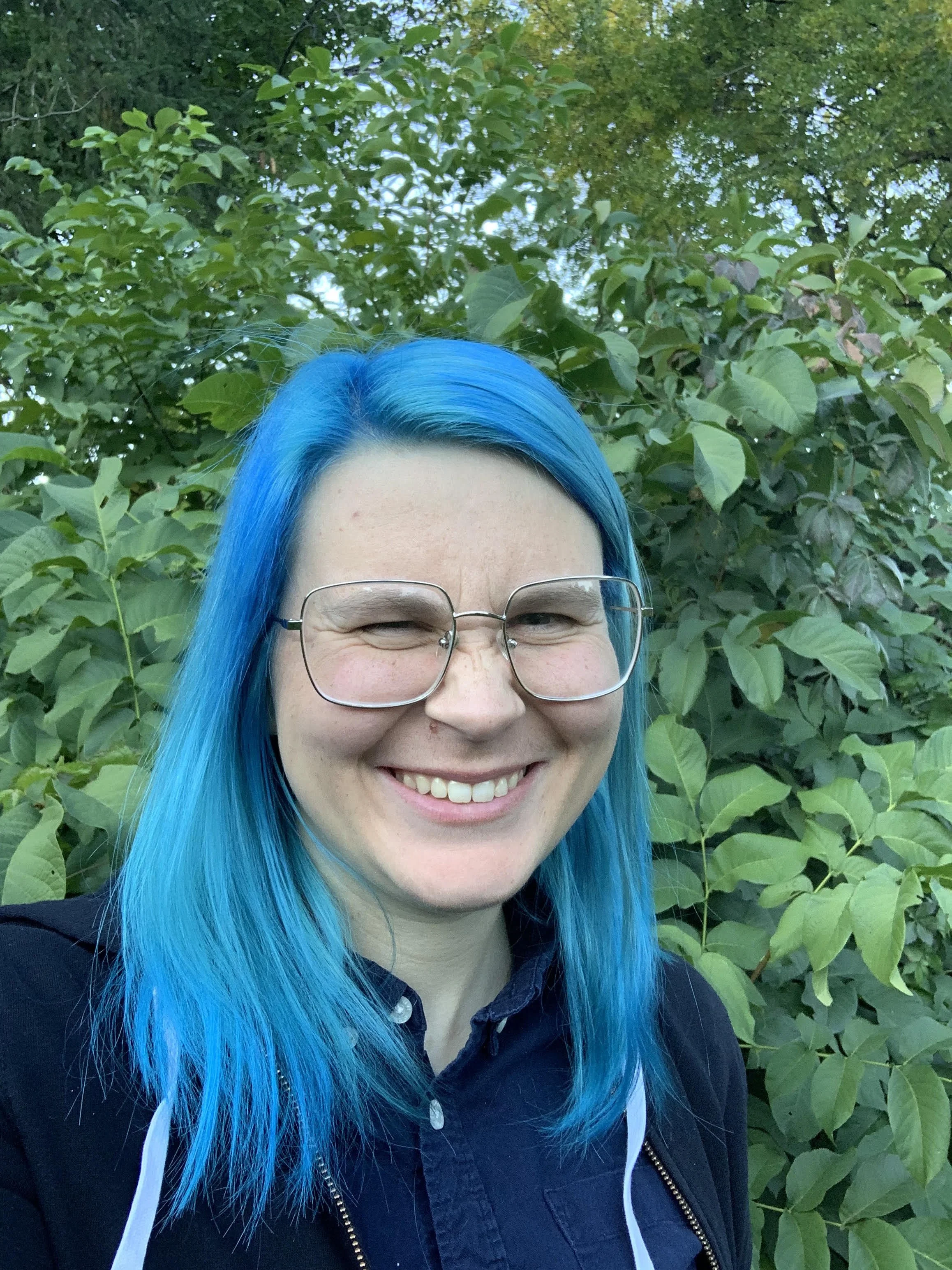 Close-up of a young woman with long blue hair smiling at the camera against a green leafy background.