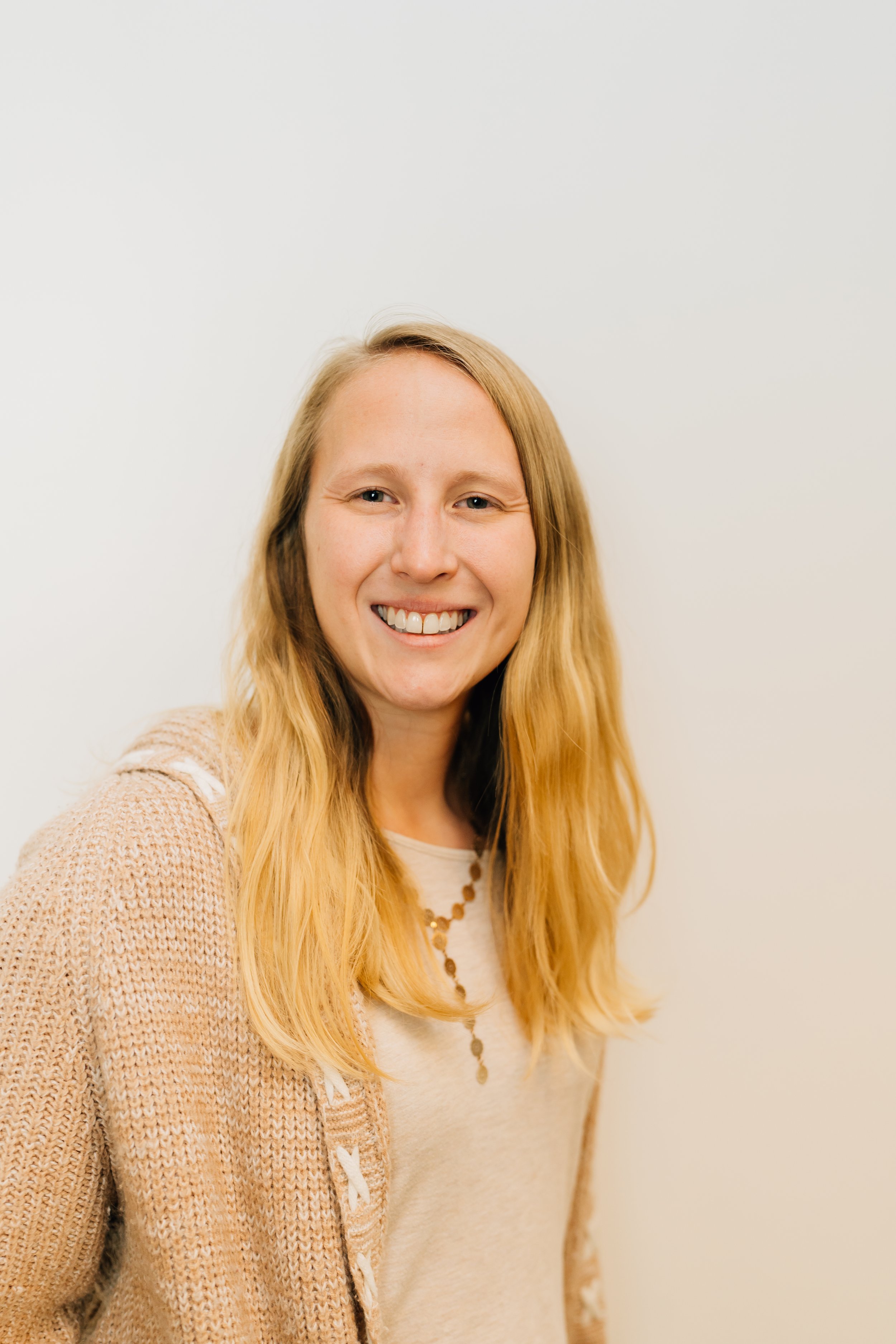 Close-up of a young woman with long blonde hair smiling at the camera against a plain white wall.