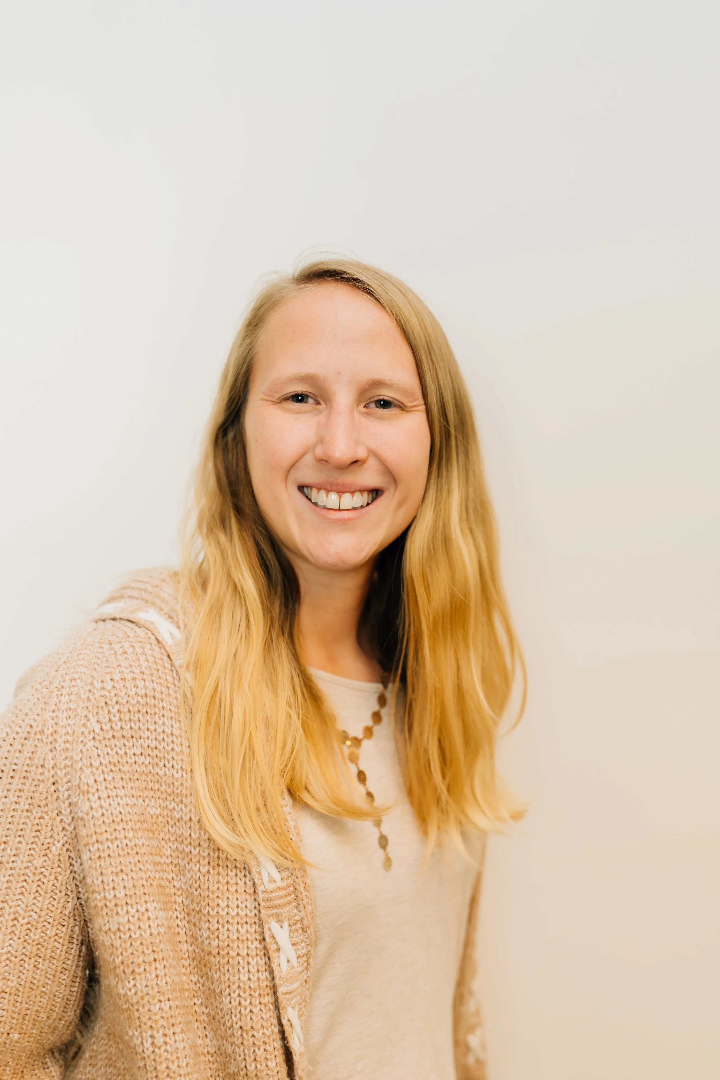 A woman with long blonde hair smiling at the camera, wearing a beige knitted cardigan and a cream-colored top, standing against a plain white background.