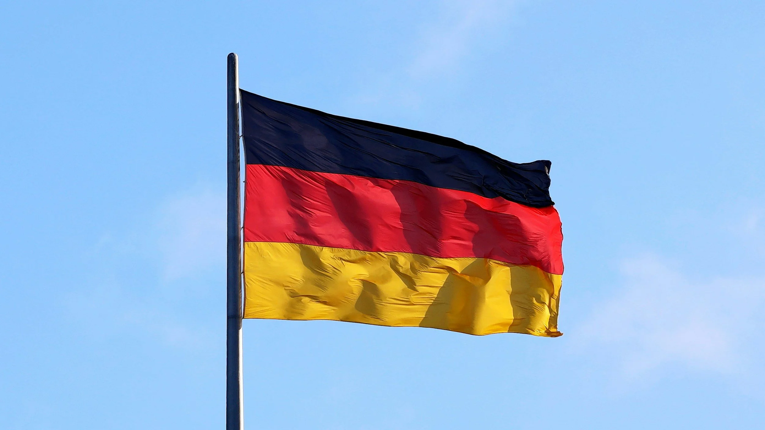 The national flag of Germany flying on a pole against a clear blue sky, with horizontal black, red, and gold stripes.