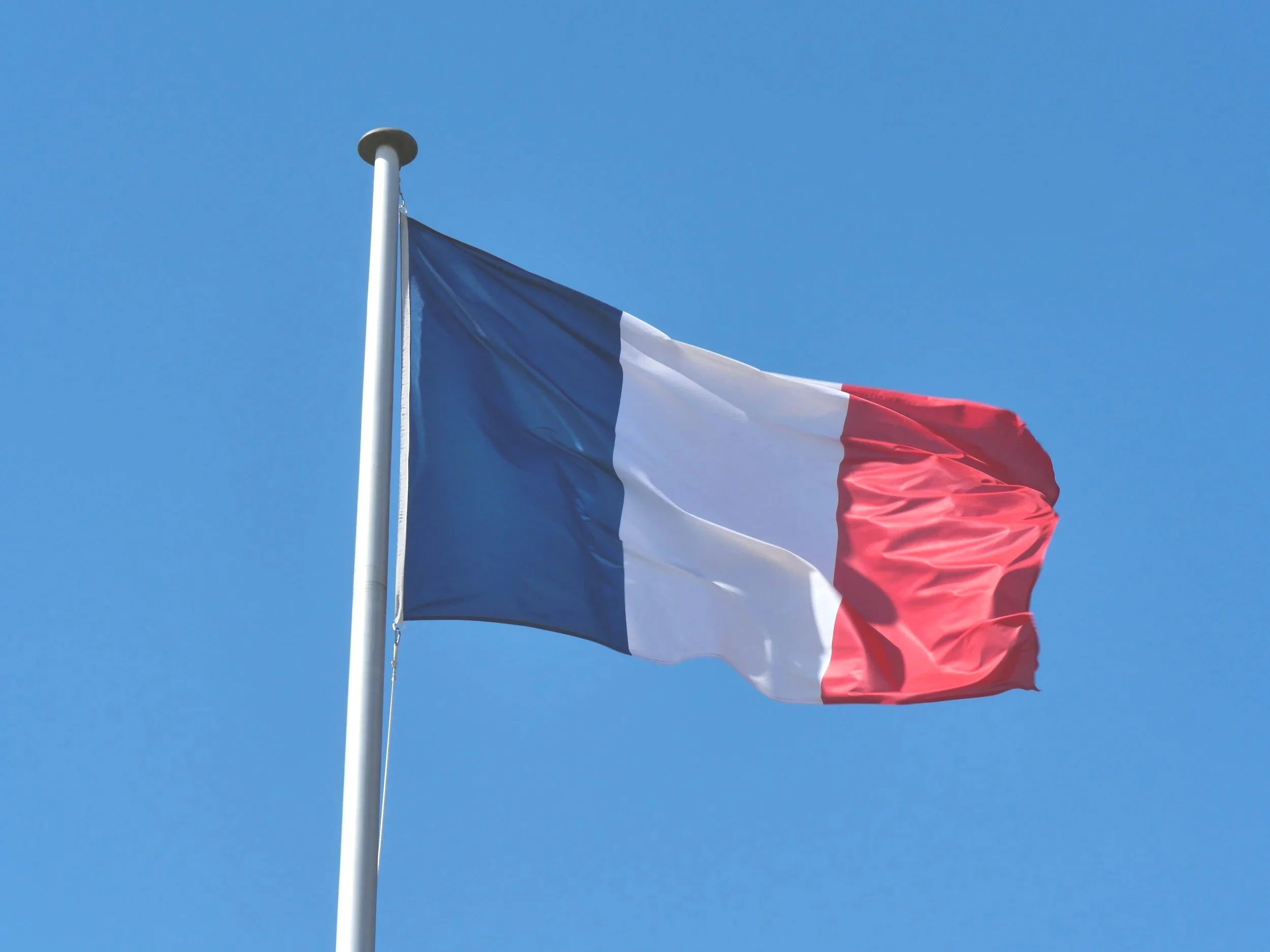 French flag with blue, white, and red vertical stripes flying against a clear blue sky.