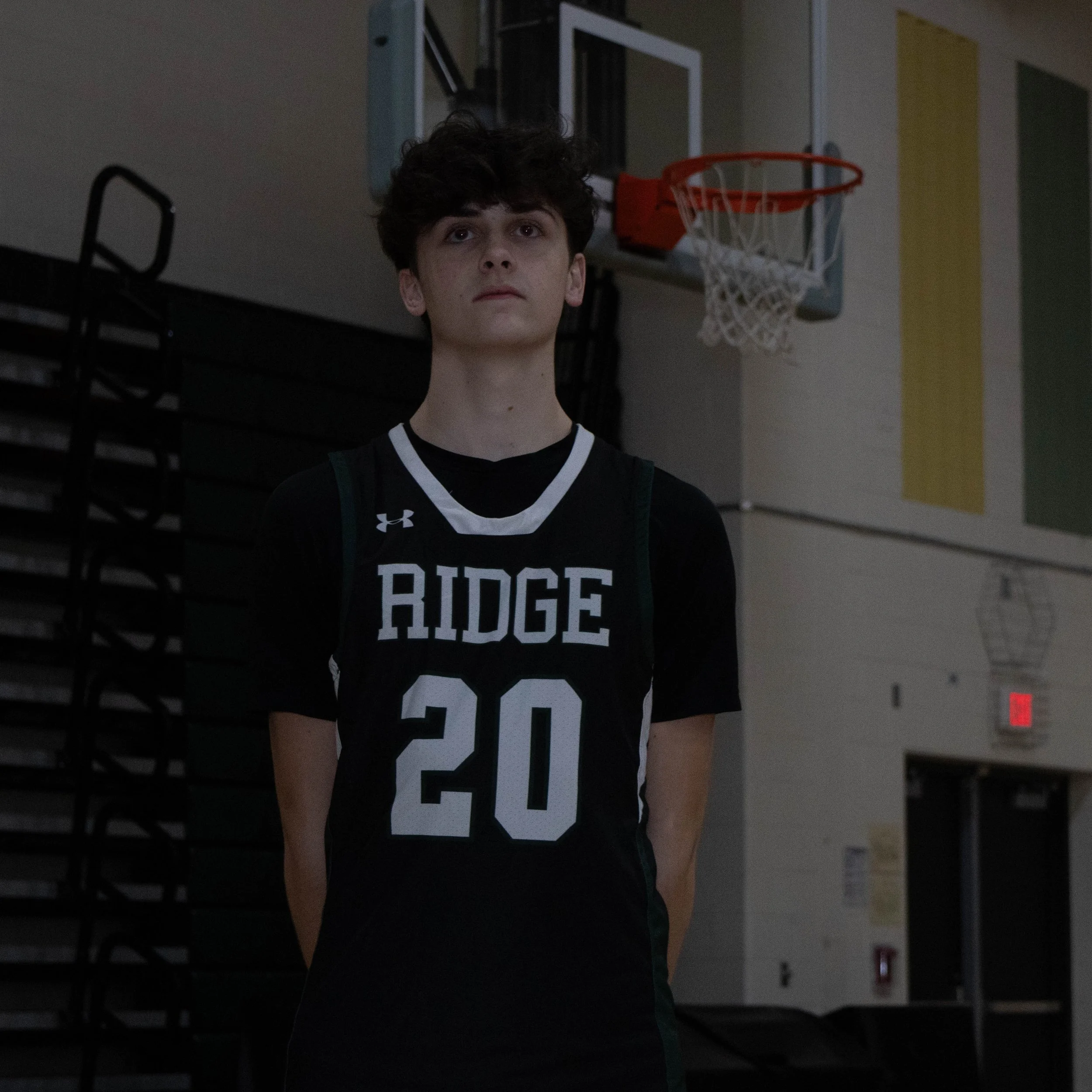 A young male basketball player standing in a gymnasium, wearing a black jersey with the word 'AIDGE' and the number 20 on it, with a basketball hoop visible in the background.