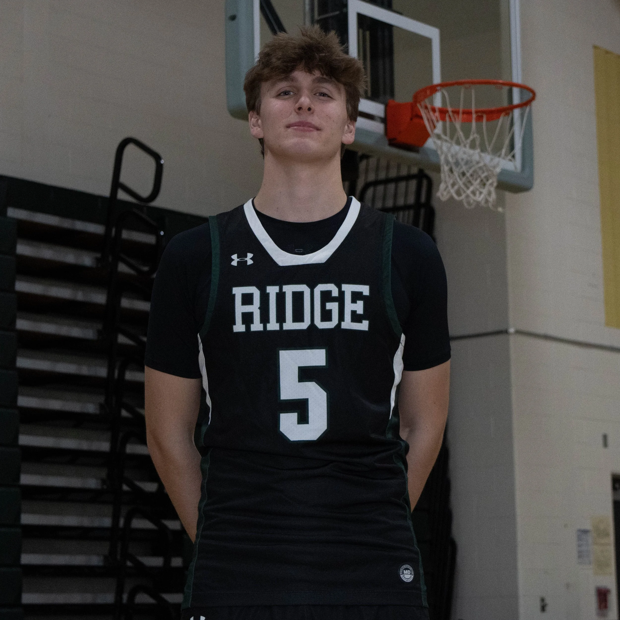 A teenage boy in a black basketball uniform with the number 5 and 'RIDGE' written on it, standing in a gymnasium with a basketball hoop in the background.