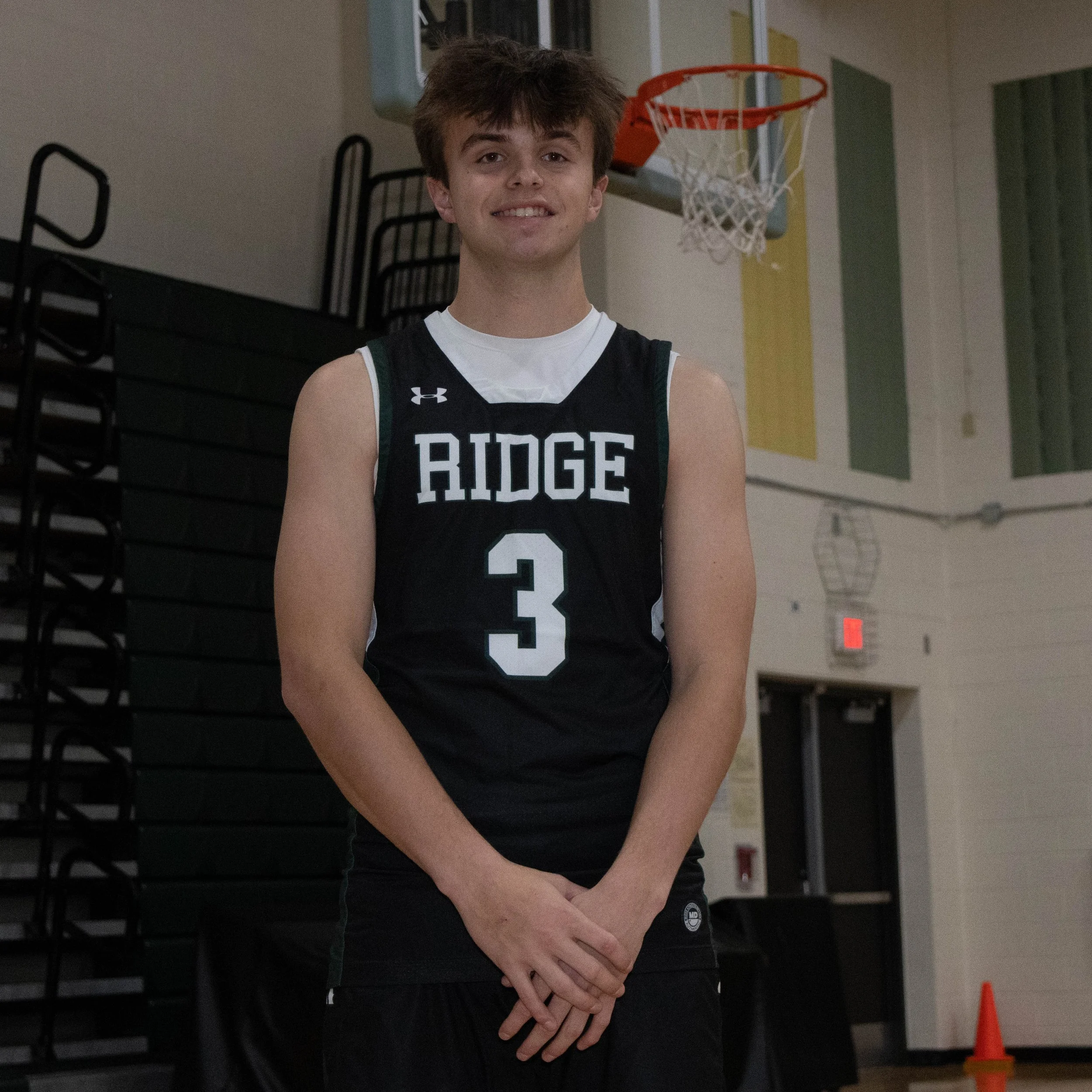 A young male basketball player stands in a gymnasium wearing a RIDGE team jersey with the number 3, smiling at the camera.
