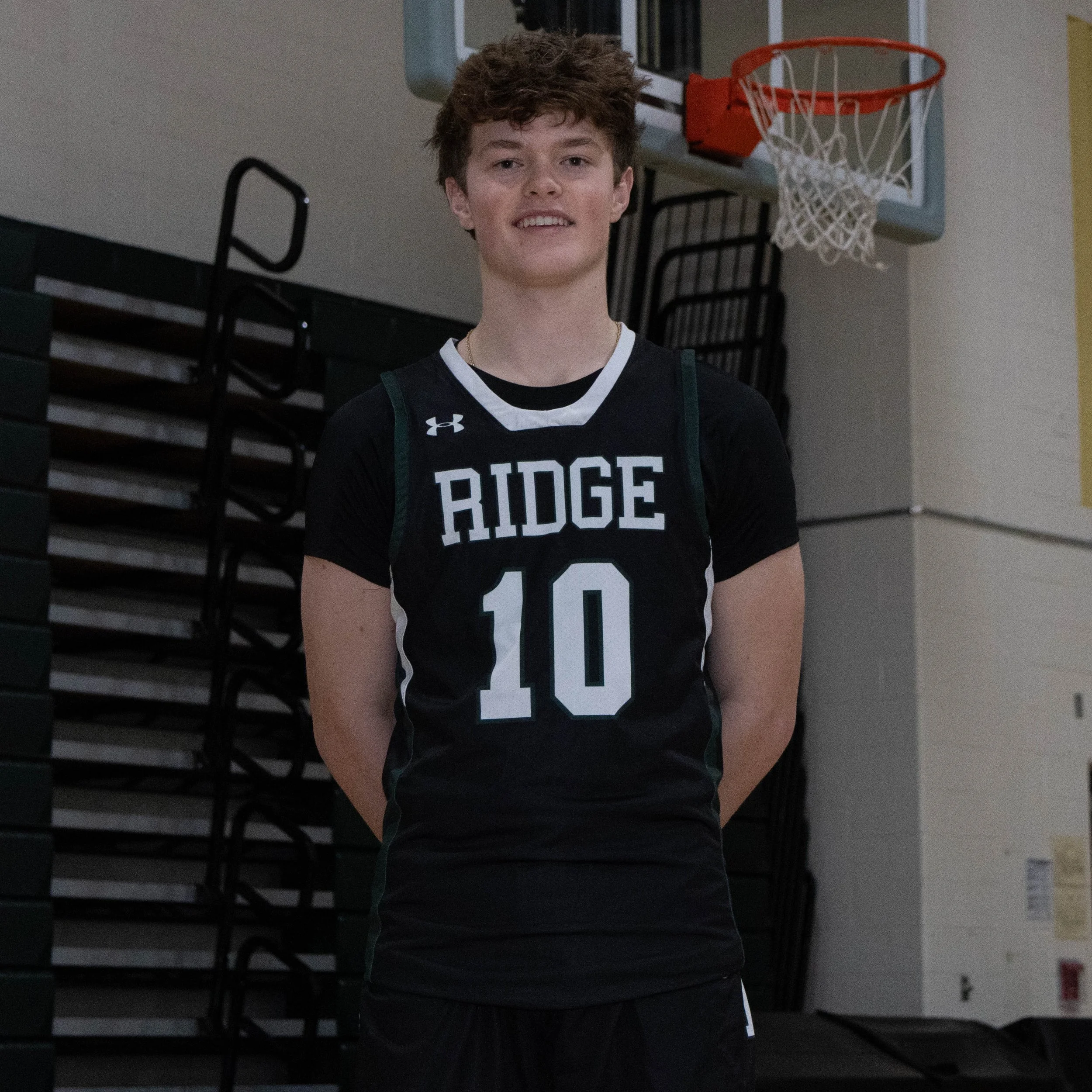 Young male basketball player standing in a gymnasium, wearing a black 'Ridge' jersey with the number 10, smiling and looking at the camera.