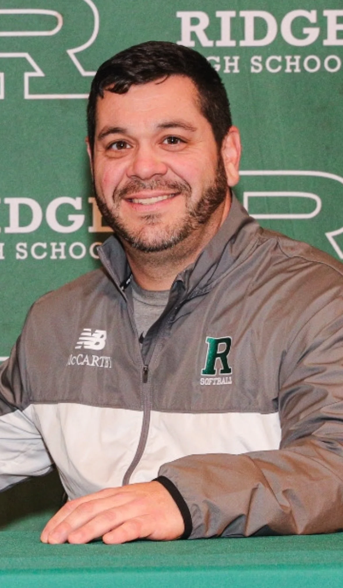 A smiling man with dark hair and a beard wearing a gray and white jacket sitting at a table in front of a green background with 'Ridge High School' printed on it.