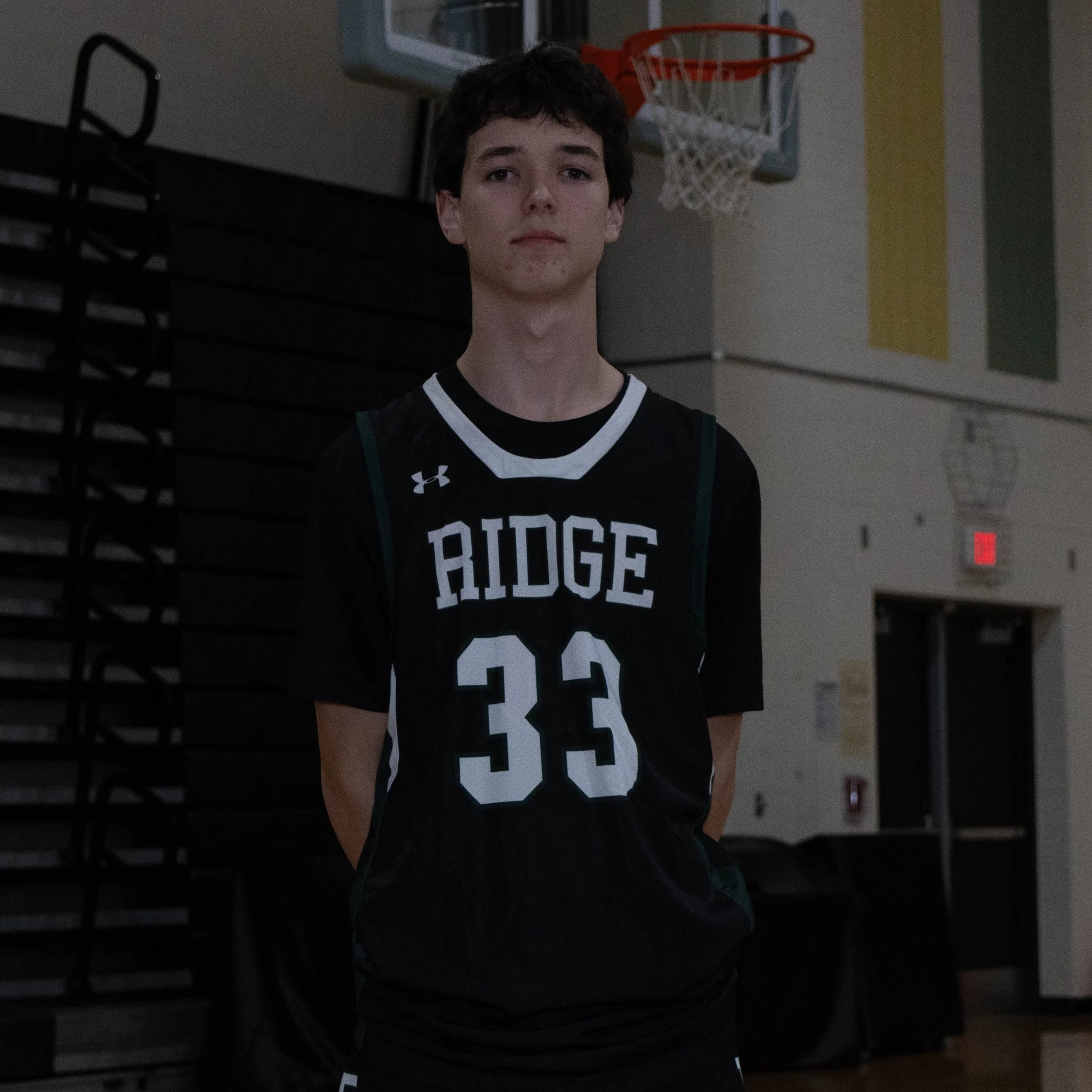 Young male basketball player in a black jersey with the words 'RIDGE' and the number 33, standing with his hands behind his back in a gymnasium.