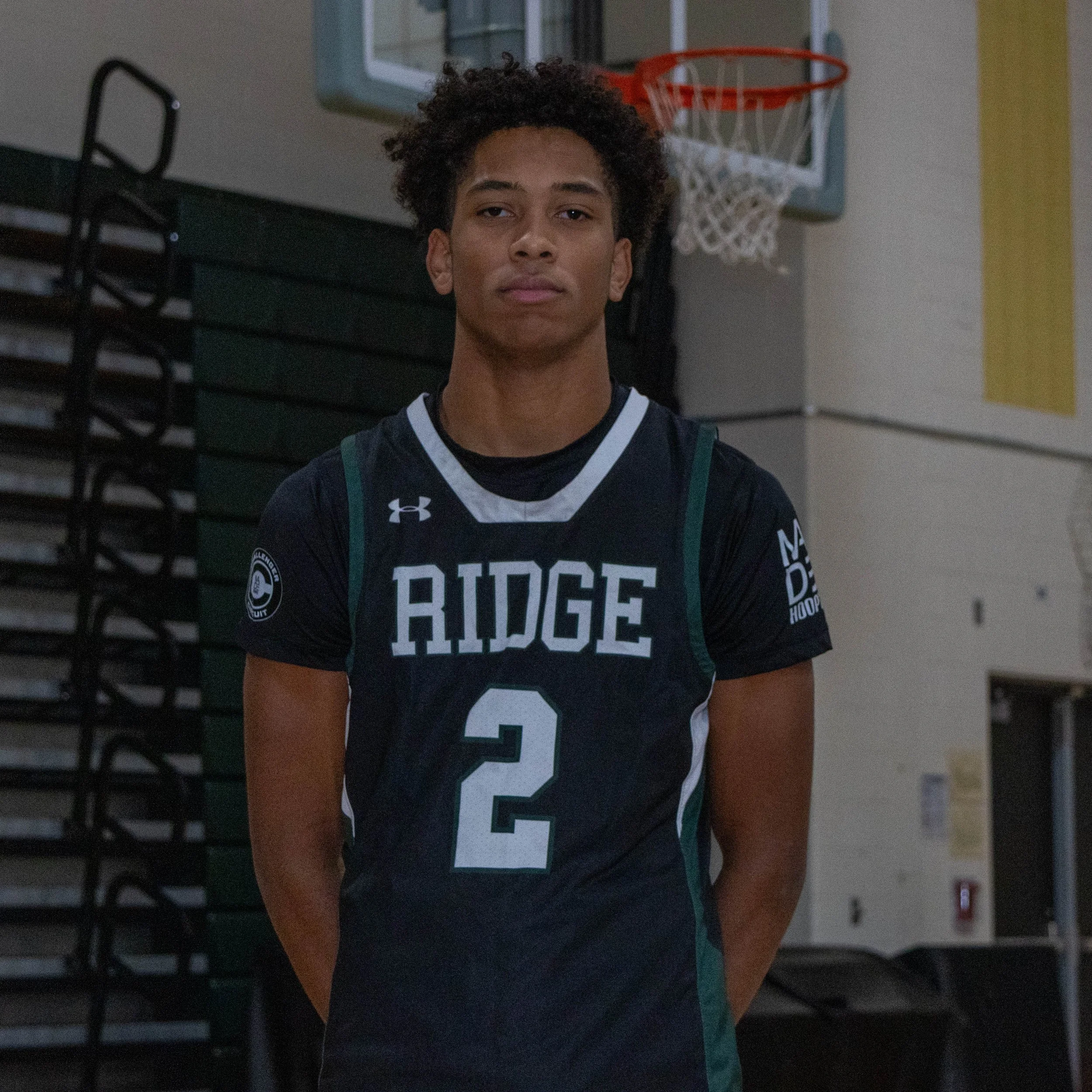 Young male basketball player standing in a gymnasium, wearing a black jersey with green and white trim that reads 'RIDGE' and the number 2.