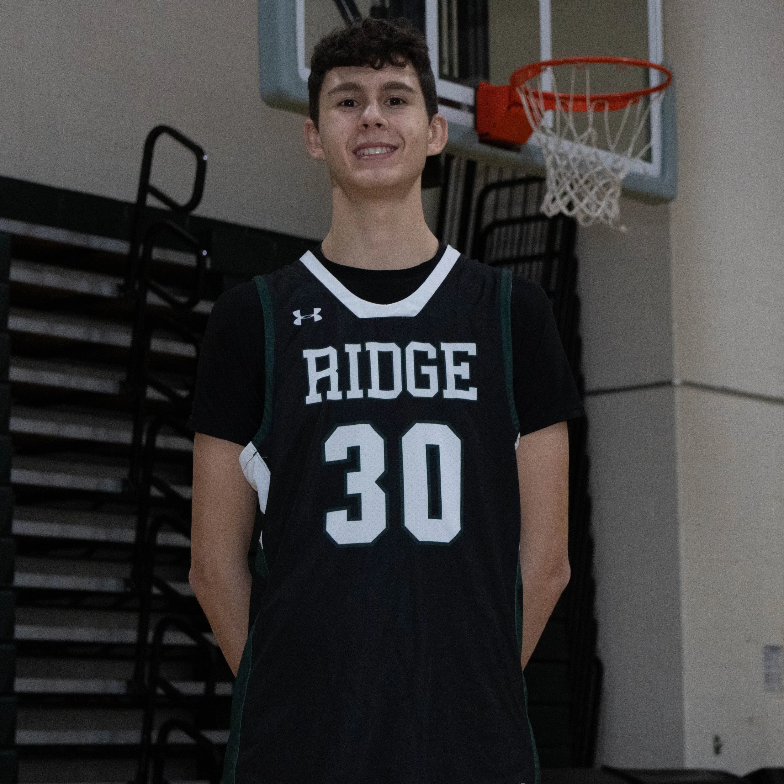 Young male basketball player posing for a photo in a gymnasium, wearing a black basketball jersey with the word 'RIDGE' and the number '30' on it, standing in front of a basketball hoop.