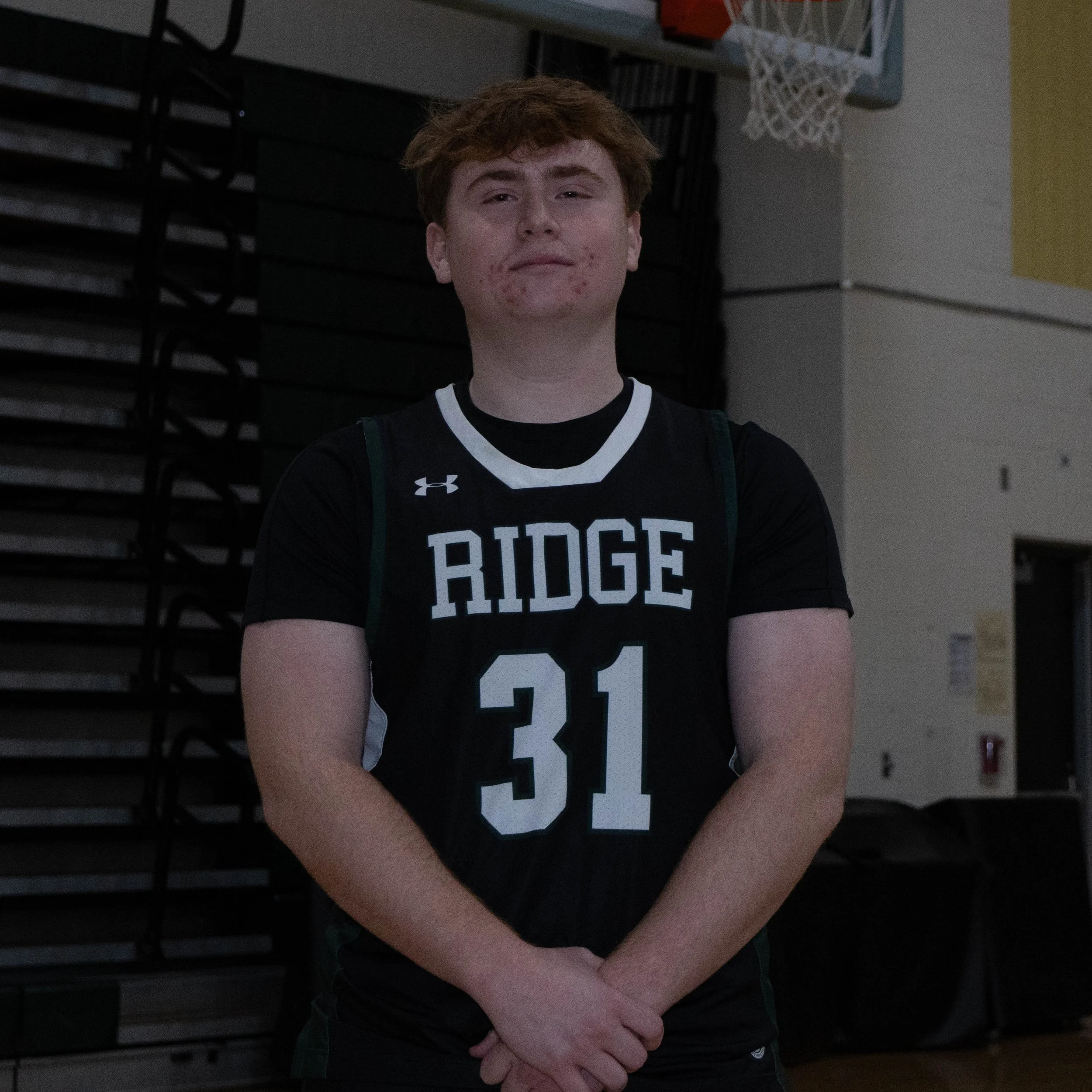 A young male athlete wearing a black basketball jersey with 'RIDGE' and the number 31, standing in a gymnasium in front of a basketball hoop, with his arms crossed.