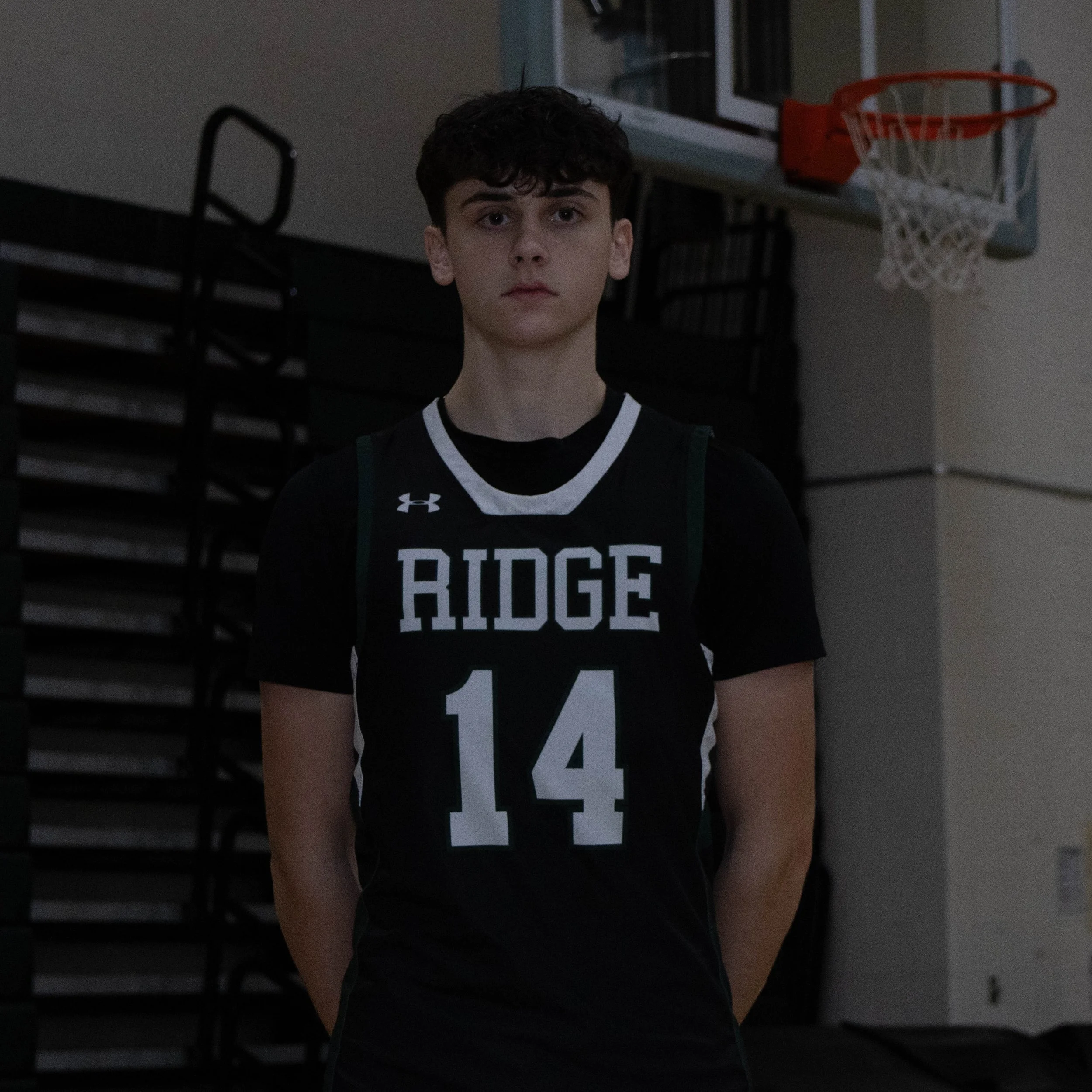 A young male basketball player in a black jersey with 'RIDGE' and the number 14, standing in a gym with a basketball hoop in the background.