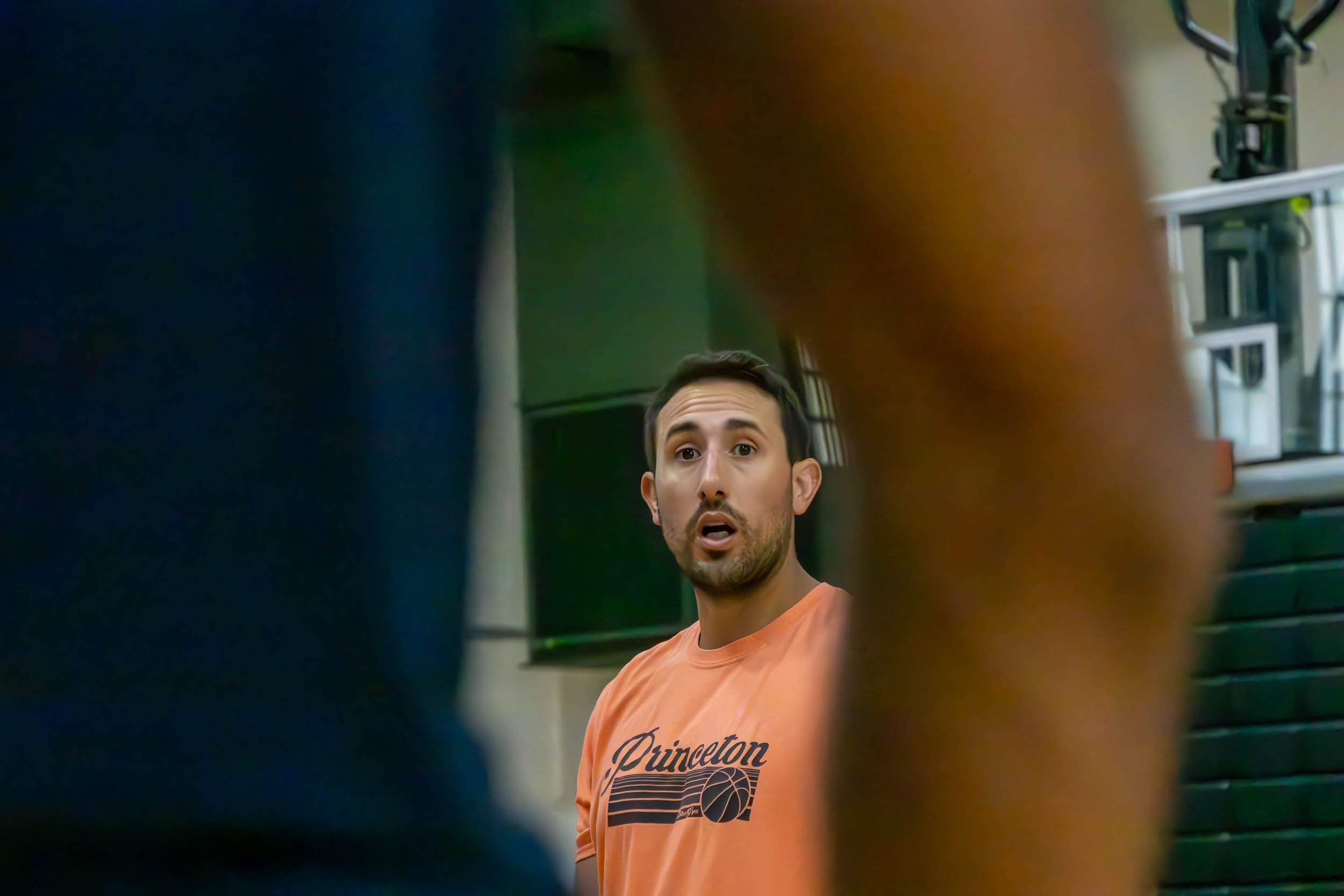 A man in an orange Princeton basketball t-shirt is looking surprised or confused, with a blurred foreground of two people standing close to him.