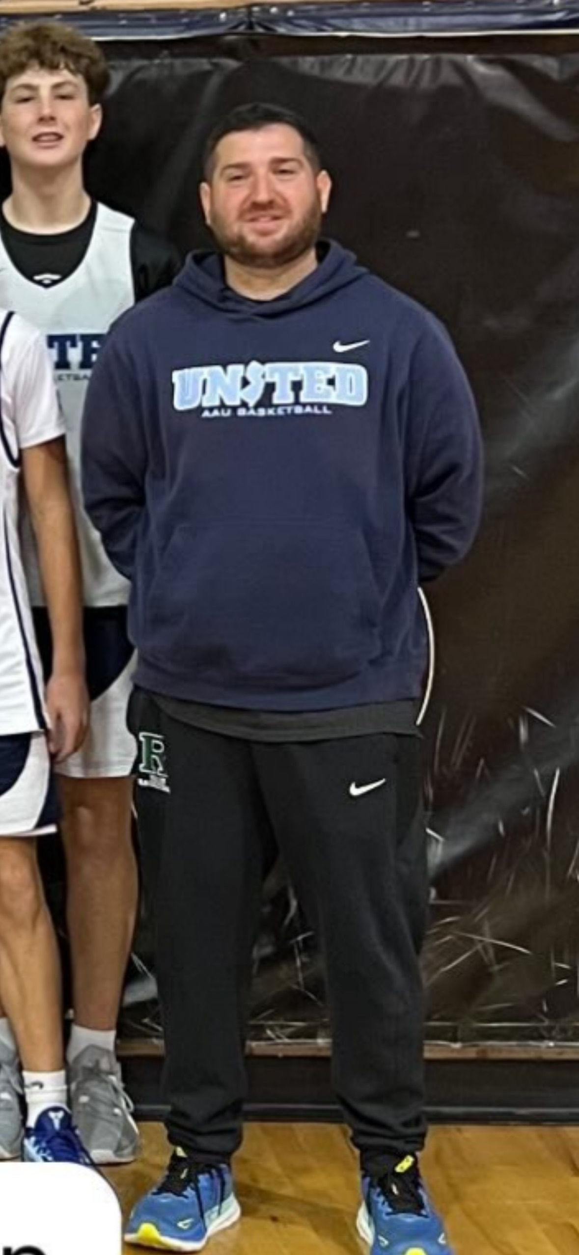 Man wearing a dark blue hoodie with 'UNTED AAU BASKETBALL' written on it, black Nike pants, and blue sneakers, standing indoors on a wooden floor in front of a black backdrop.