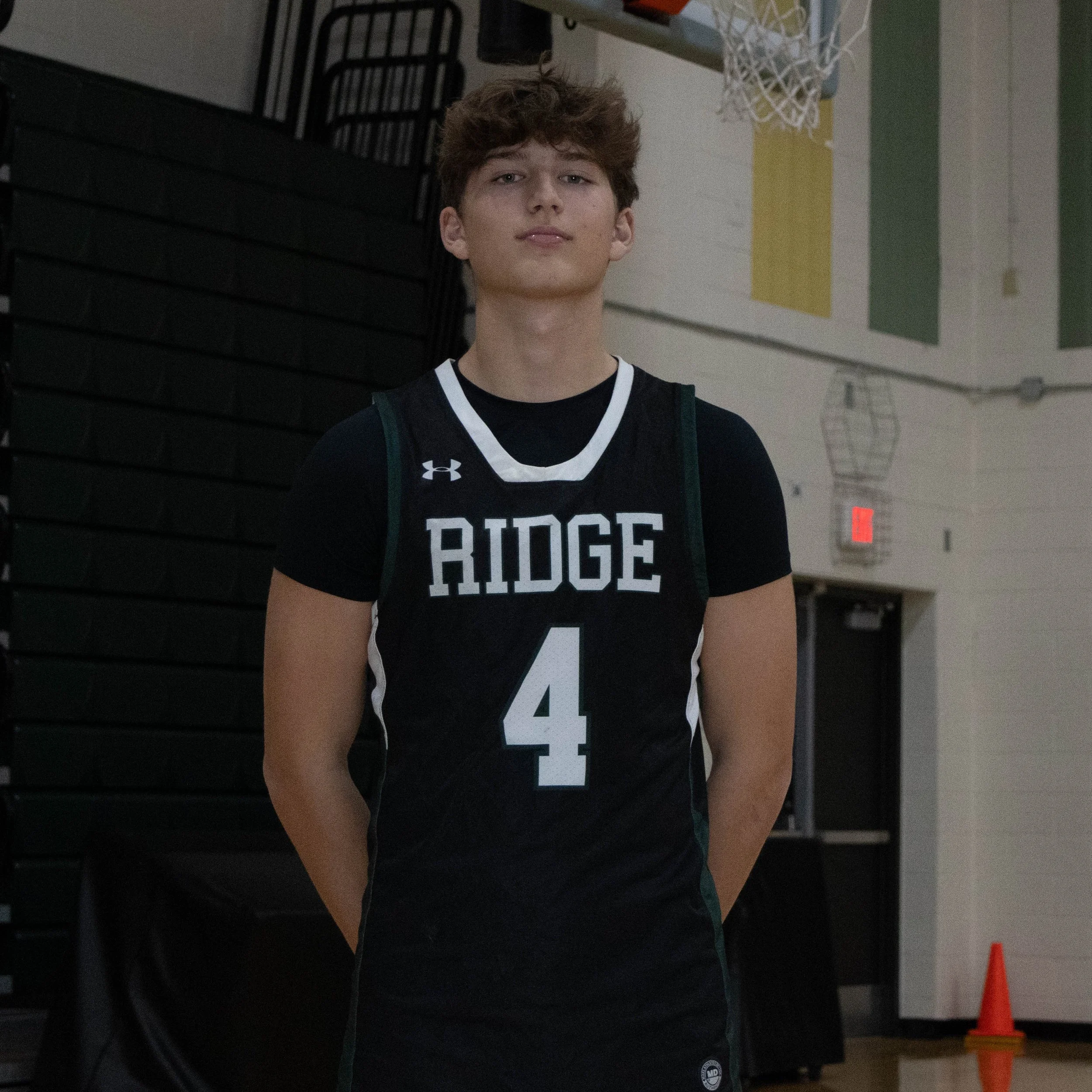A young male basketball player standing indoors, wearing a black Jersey with white and green accents, numbered 4, with 'RIDGE' written on the front.
