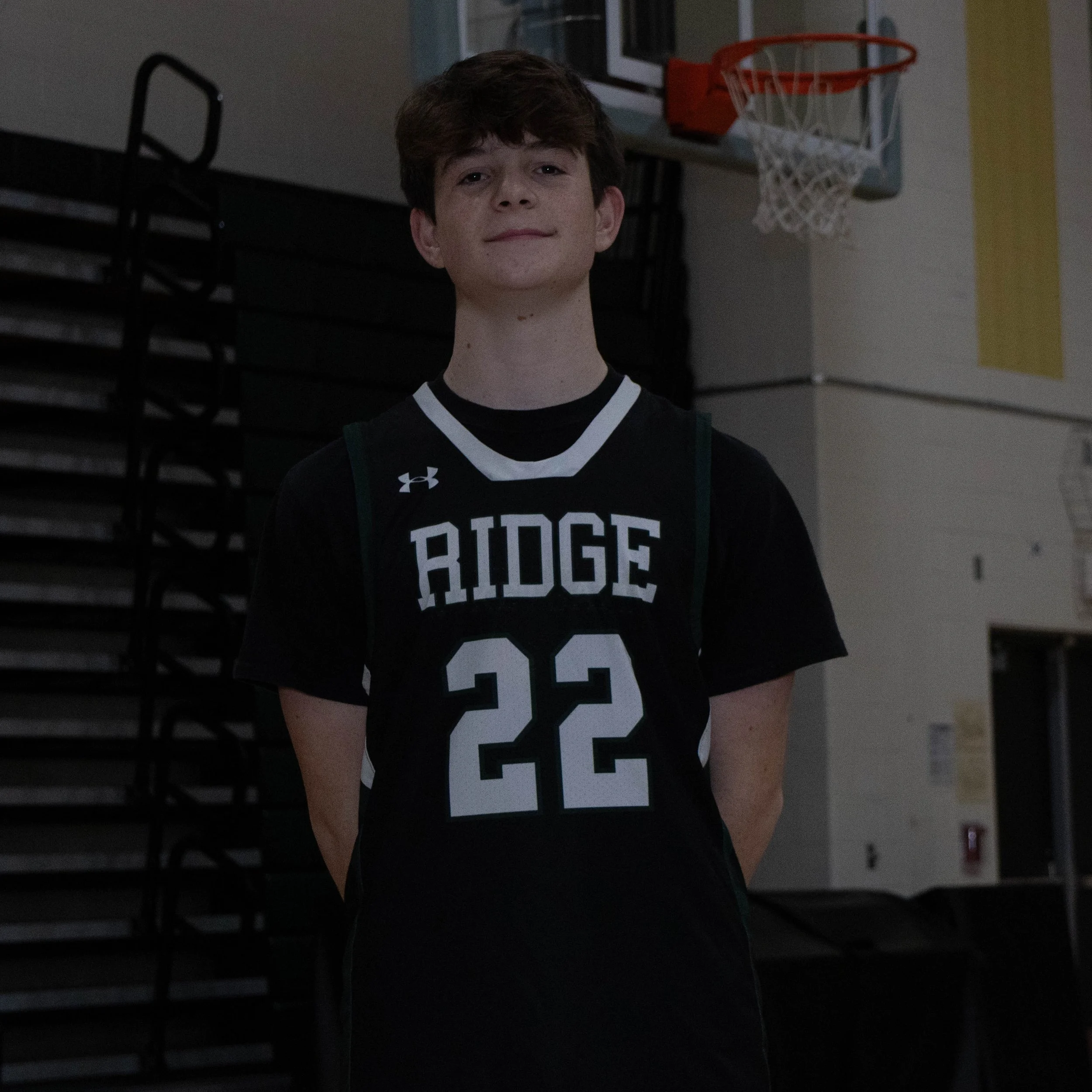 A young male basketball player in a black and white jersey with the word 'RIDGE' and the number 22, standing inside a gymnasium in front of a basketball hoop.