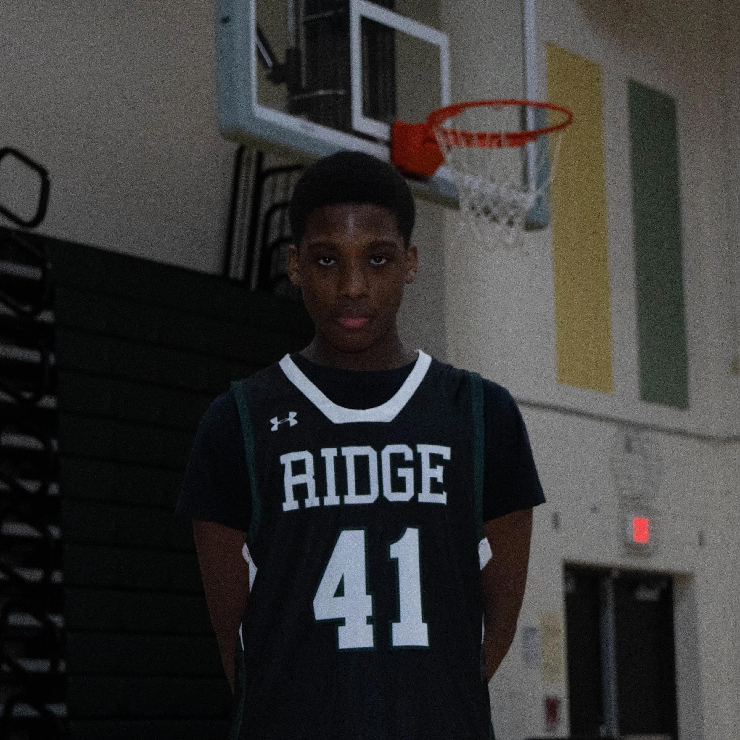 A young African American boy wearing a black basketball jersey with the number 41 and the word 'Ridge' on it, standing in a gymnasium in front of a basketball hoop.