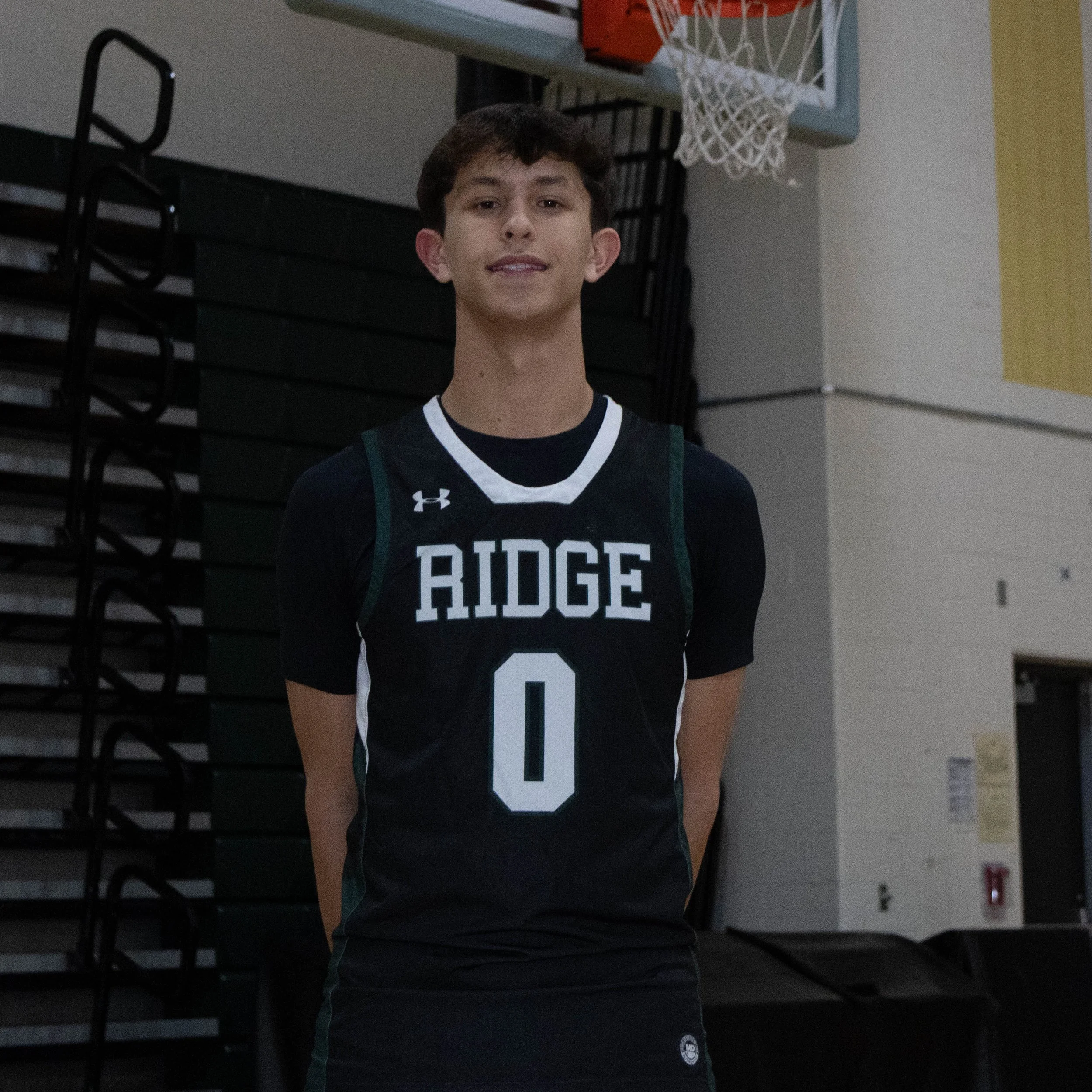 Young male basketball player wearing a black jersey with 'RIDGE' and the number 0, standing in a gymnasium near a basketball hoop.