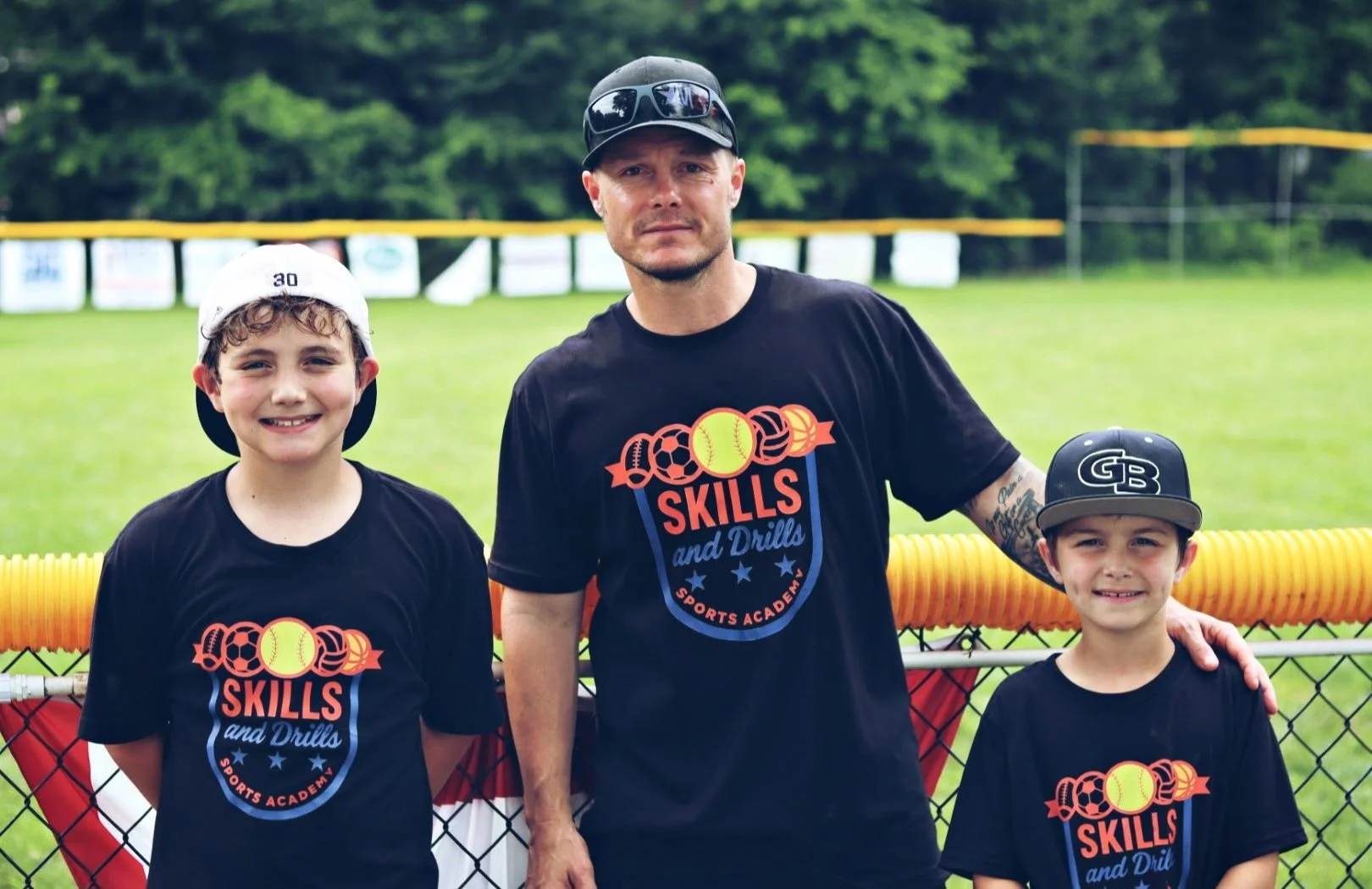 Three people standing on a baseball field, wearing black t-shirts with a sports skills logo, with a fence and trees in the background.