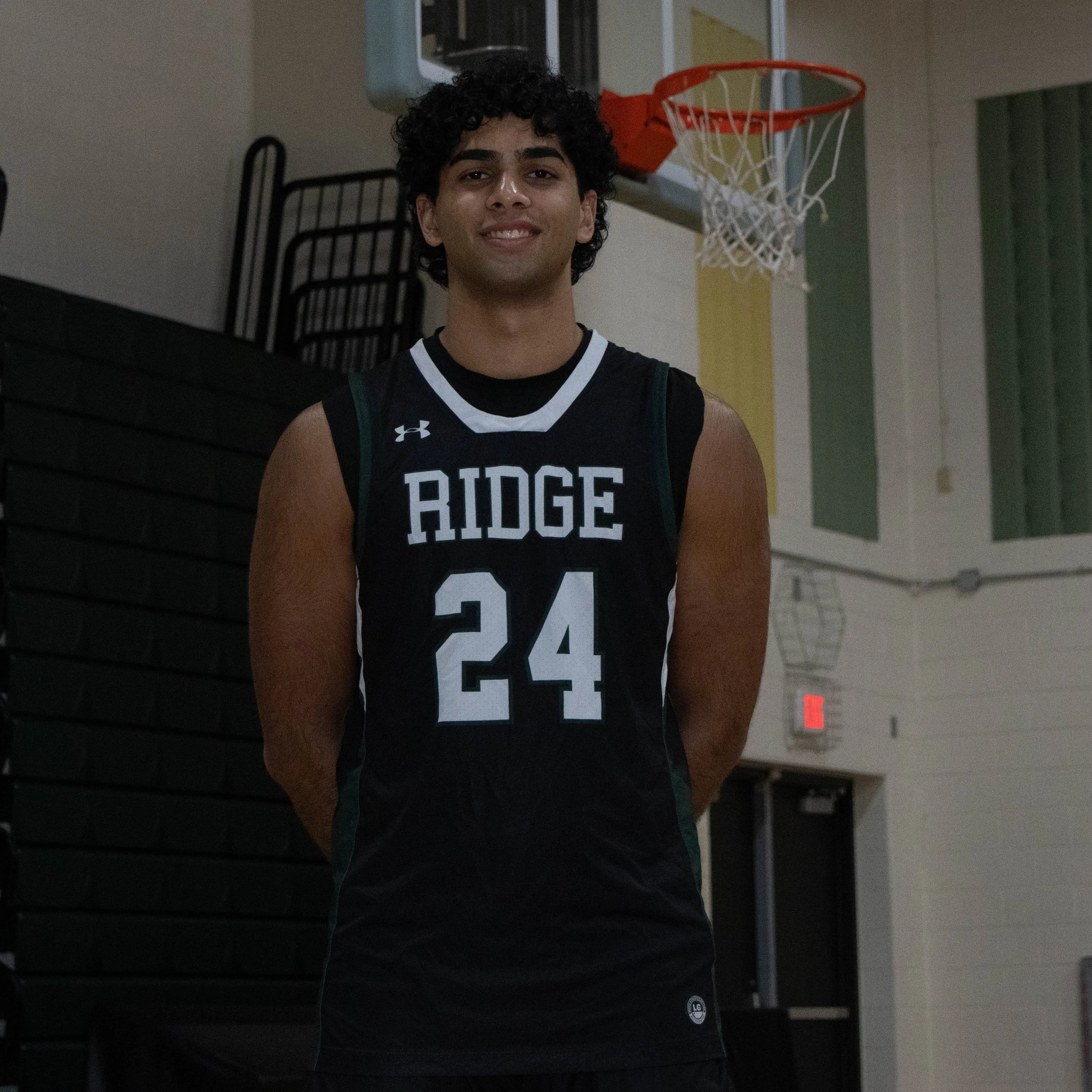 Young man in a basketball uniform standing in a gymnasium with a basketball hoop in the background.