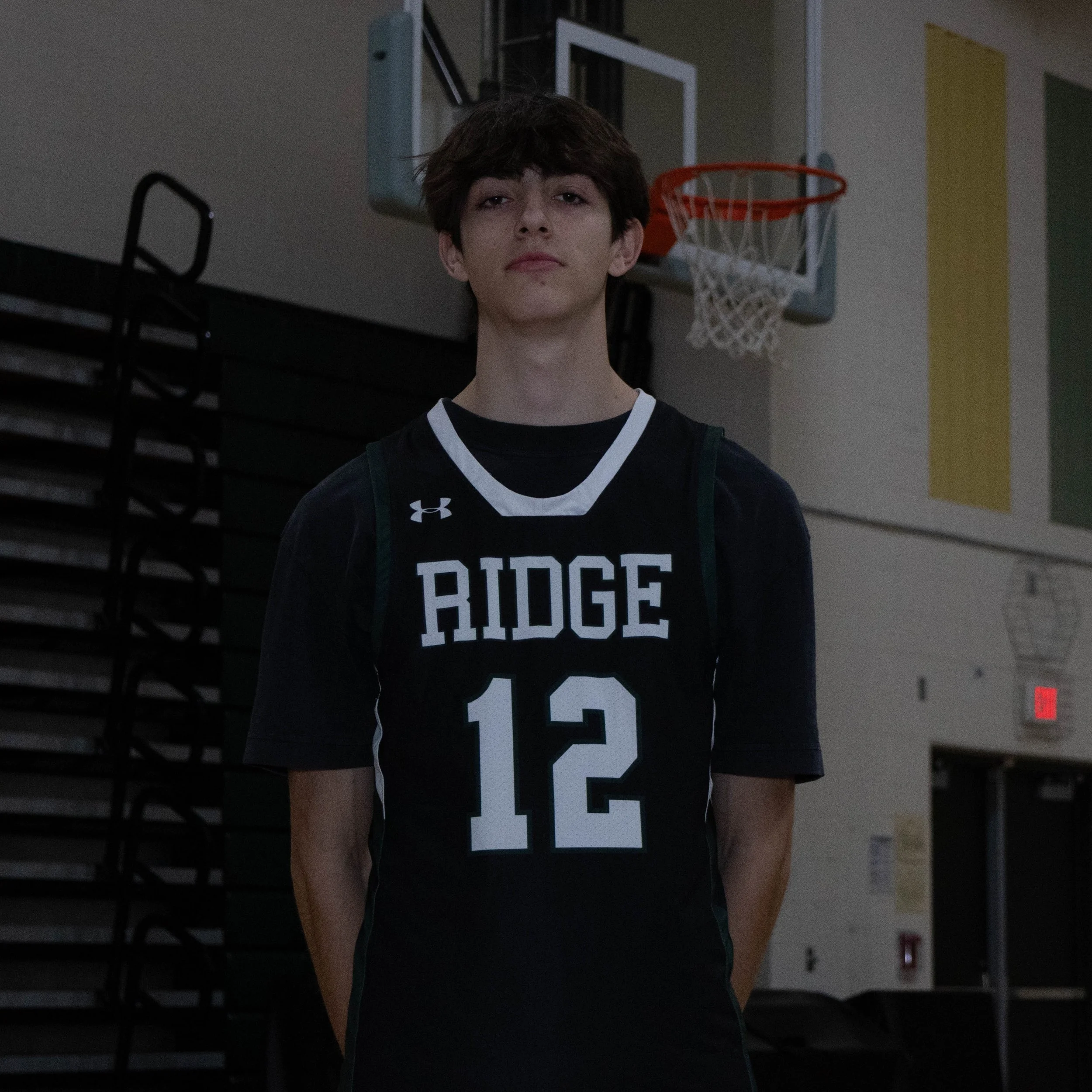A young man in a basketball uniform with the number 12 and 'Ridge' on the front, standing in a gymnasium with a basketball hoop in the background.