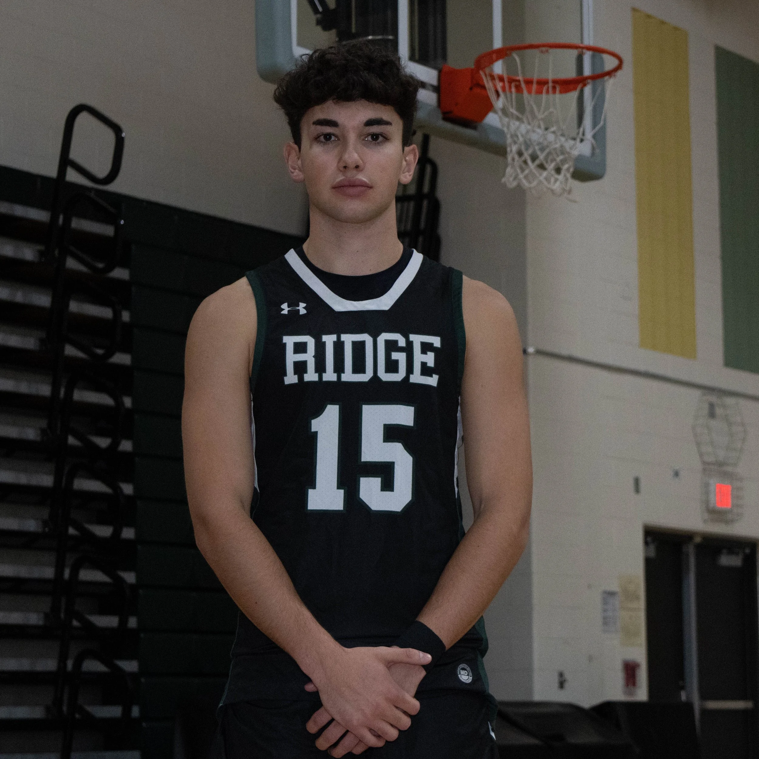 A young male basketball player stands in a gymnasium wearing a black jersey with the words "RIDGE" and the number "15" on it. He has short curly hair and is looking at the camera with a neutral expression.