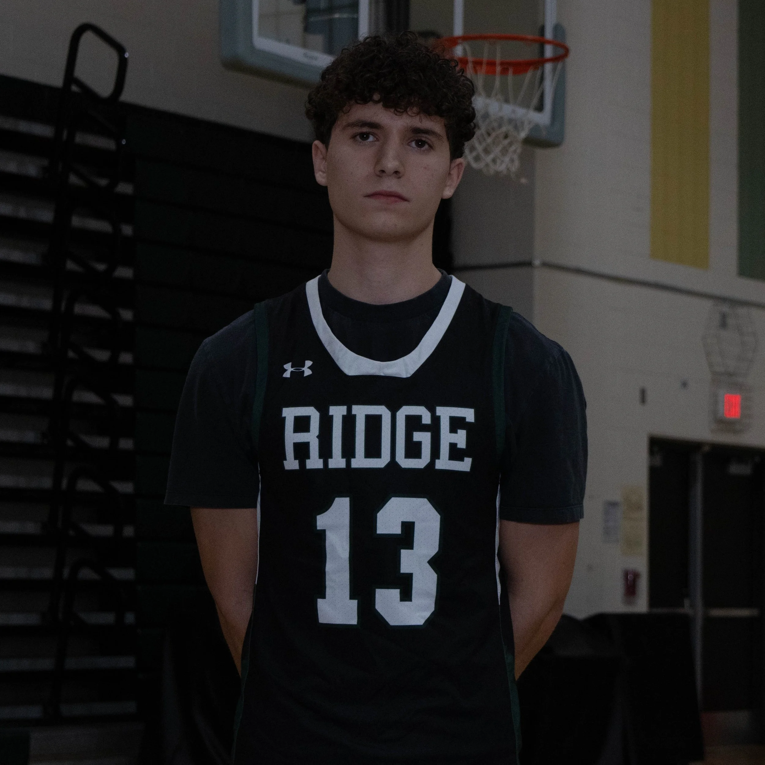 Young male basketball player in a black and white jersey with 'RIDGE' and the number 13, standing indoors in front of a basketball hoop.