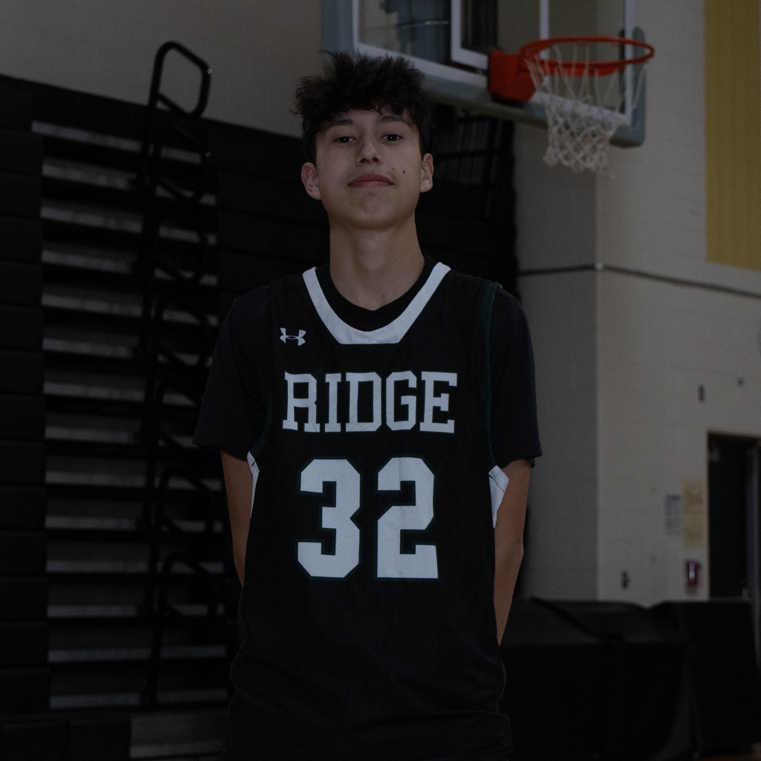 A young boy in a black basketball jersey with 'RIDGE' and the number 32, standing in a gymnasium with a basketball hoop behind him.