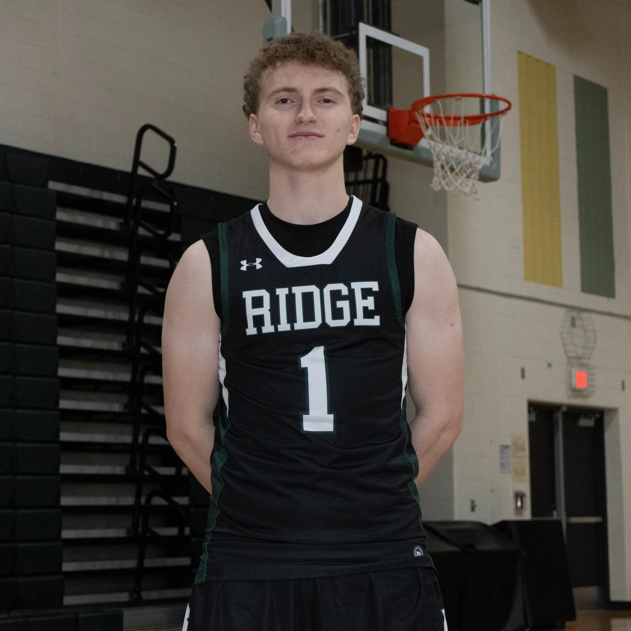 Young male basketball player standing in gymnasium wearing a black jersey with 'RIDGE' and number 1, with a basketball hoop in the background.