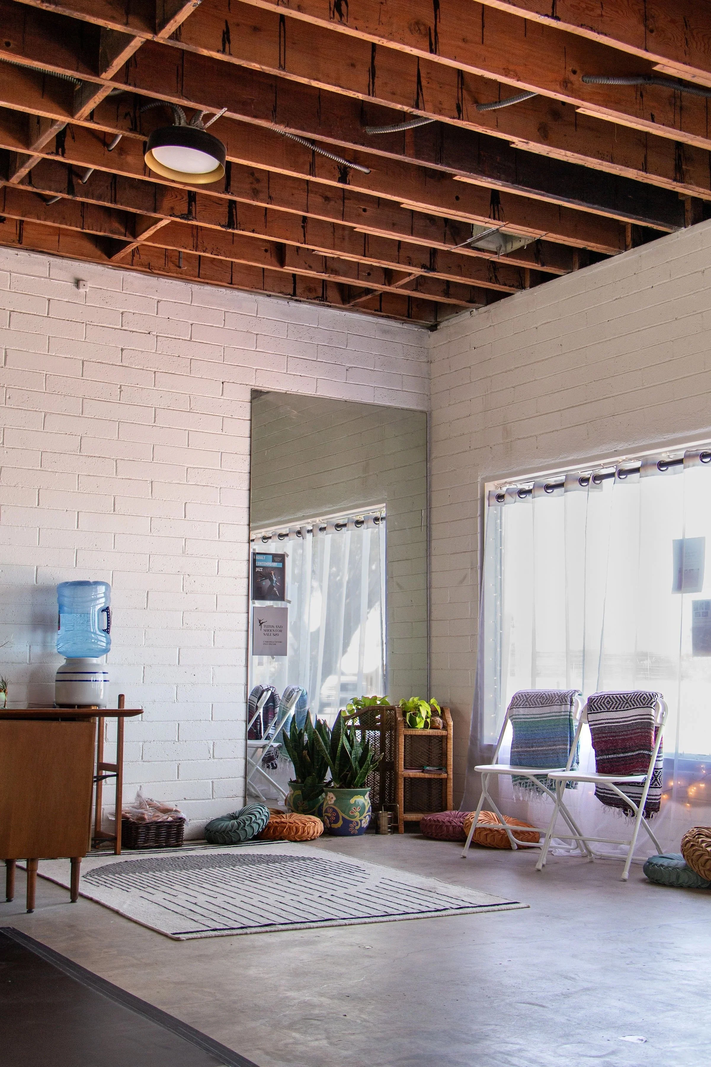 A cozy indoor space with white brick walls, a large mirror, a water dispenser, potted plants, a wicker shelf, and two white chairs with colorful woven textiles, illuminated by natural light through sheer curtains.