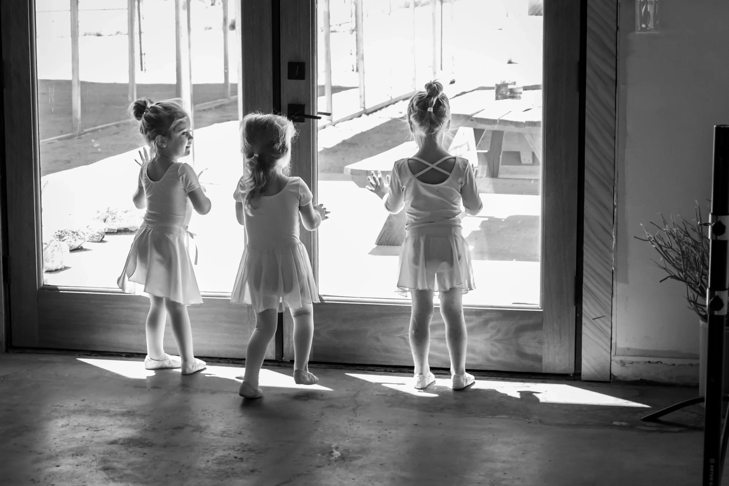 Four young girls in ballet dresses standing by a glass door, looking outside, with sunlight streaming in.