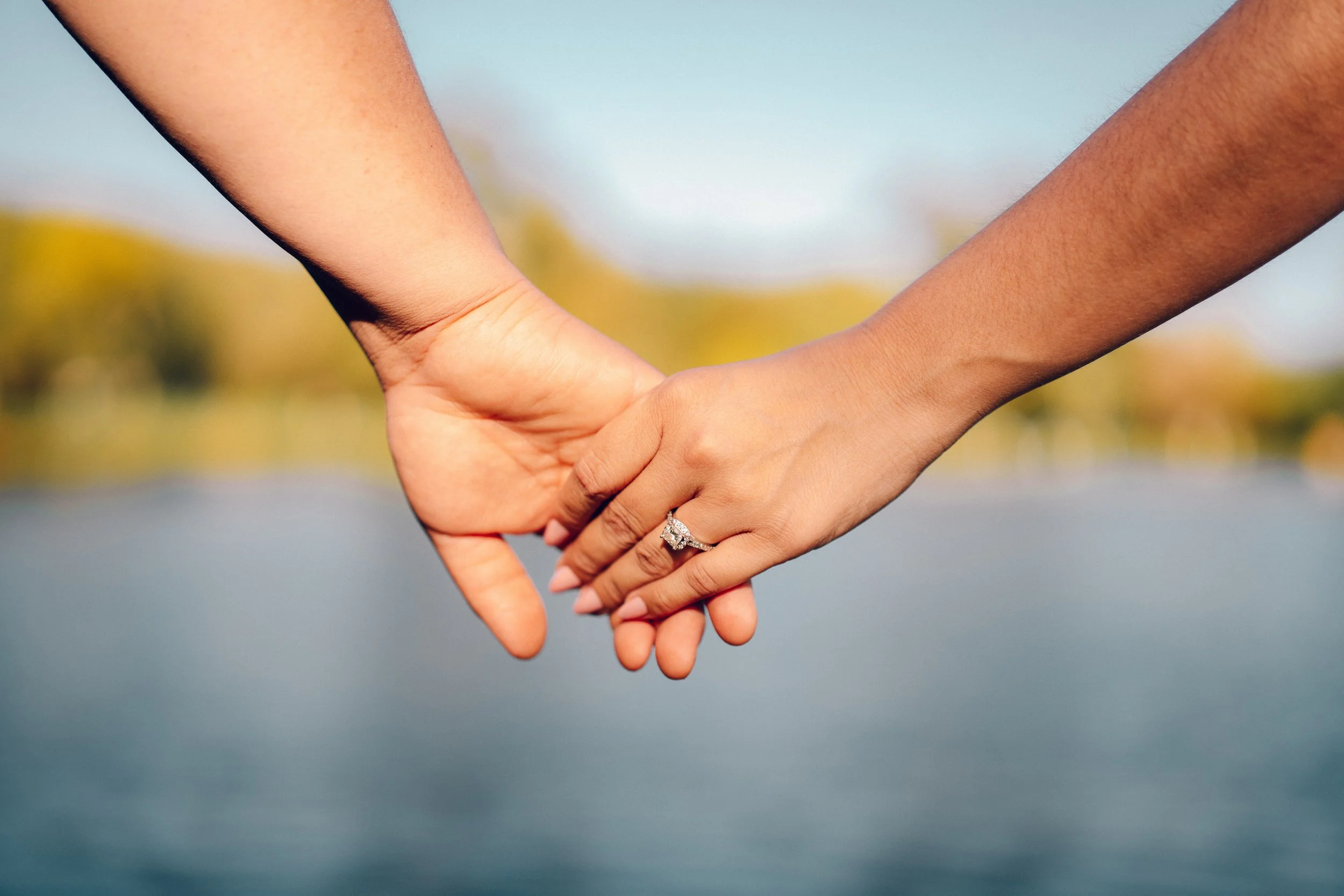 Close-up of two hands holding each other, one displaying an engagement ring, with a blurred background of outdoor scenery.