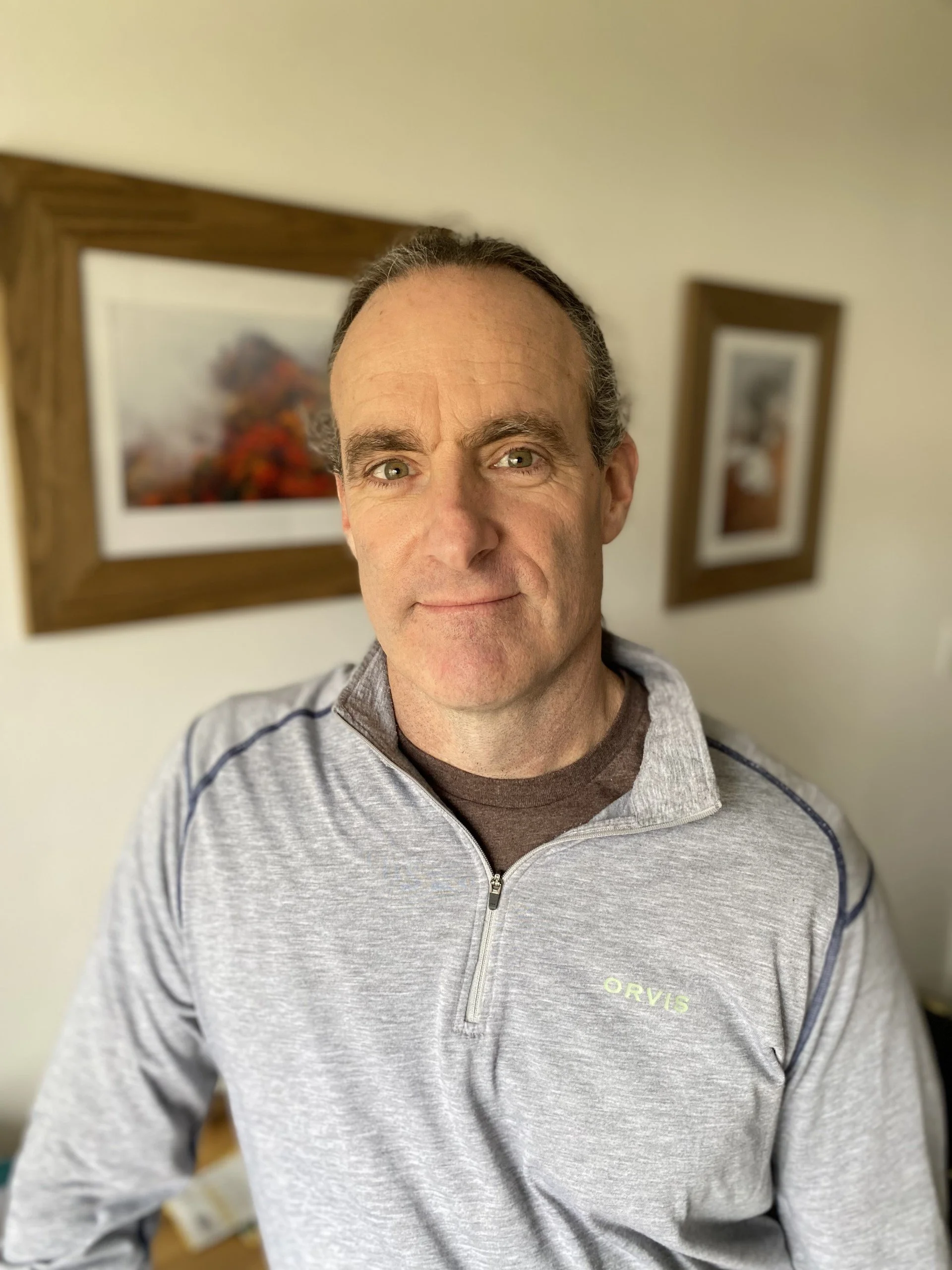A middle-aged man with short dark hair and blue eyes, wearing a light gray zip-up sports jacket with dark piping and a brown shirt underneath, standing indoors in front of a wall with two framed landscape paintings.