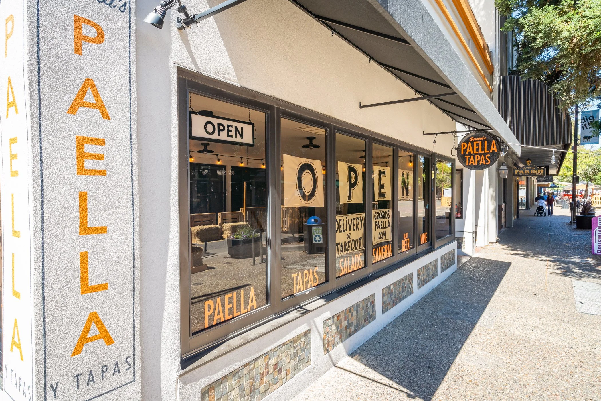 Outside view of a restaurant with large glass windows displaying signs that say "Open" and offer delivery or takeout, salads, tapas, paella, gambas, wine, and beer. A vertical sign on the wall reads "PAELLA" in large orange letters. The street is sun