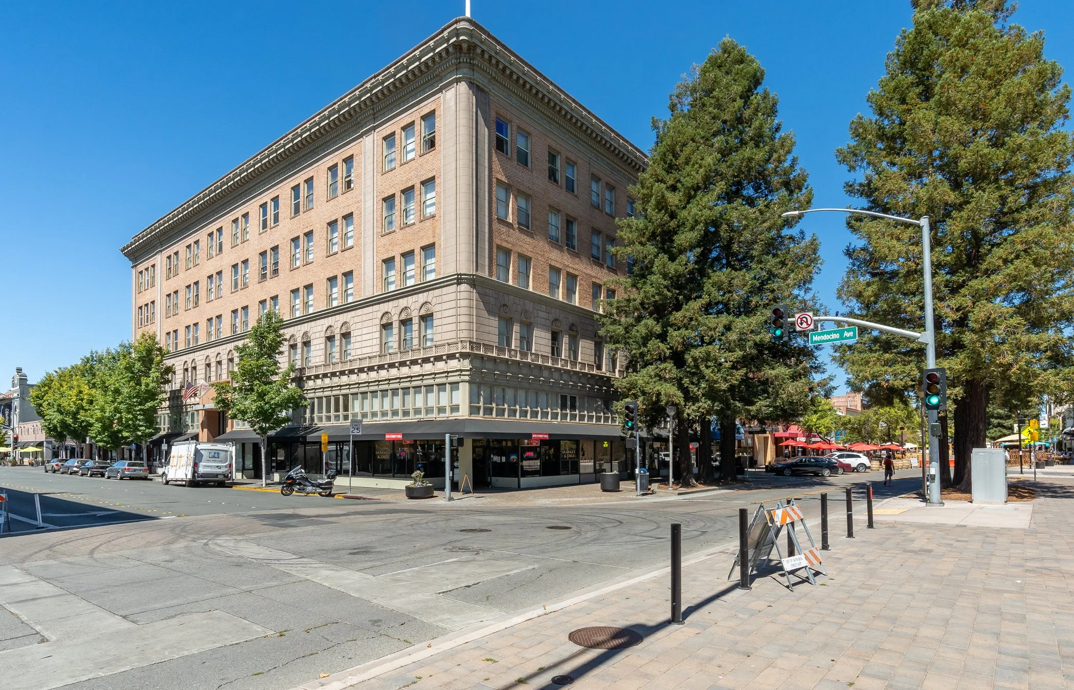 A multi-story brick building with large windows on a city corner, next to tall evergreen trees, with street signs, traffic lights, and parked cars on a clear day.