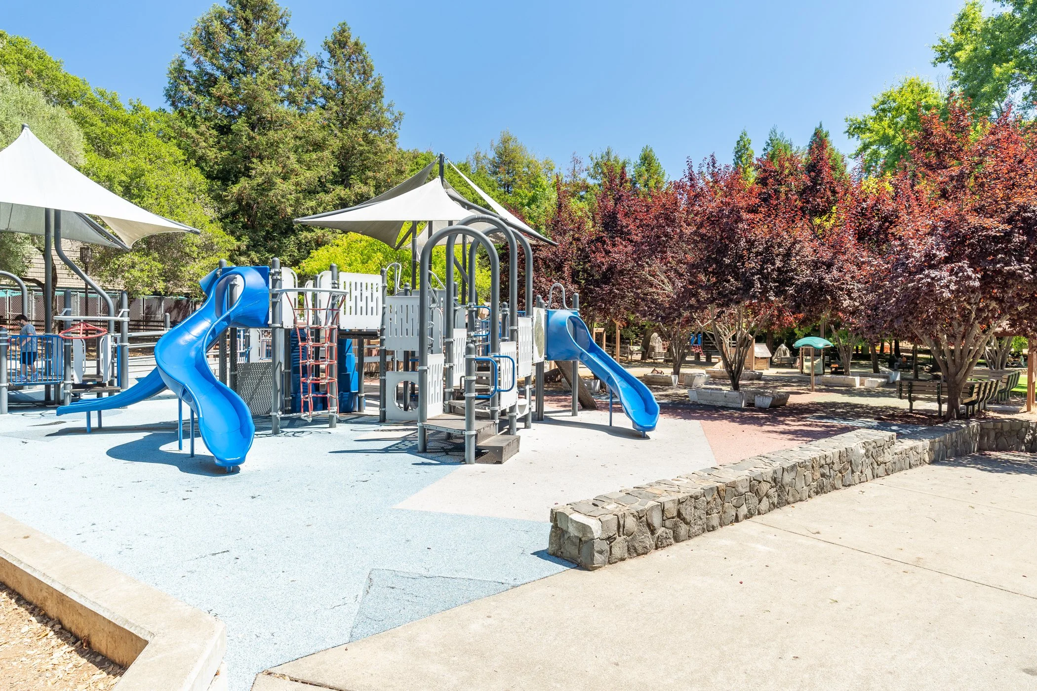 Colorful playground with blue slides, shade structures, trees with red and green leaves, and a paved pathway in a park setting.