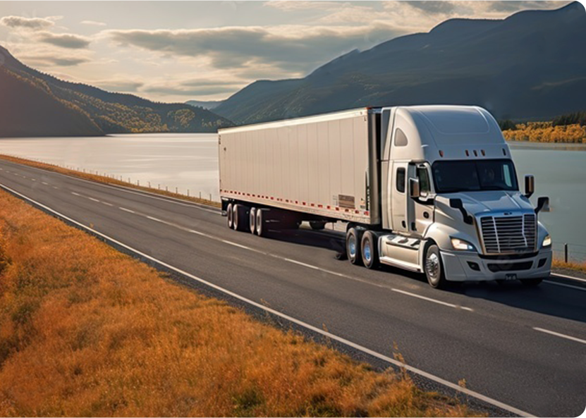 A white semi-truck driving on a highway alongside a scenic lake with mountains in the background during sunset.