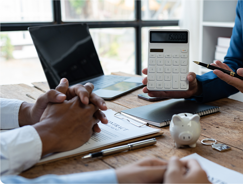 People in a business meeting at a wooden table with a laptop, notepad, piggy bank, and large calculator showing '1500000' on display.