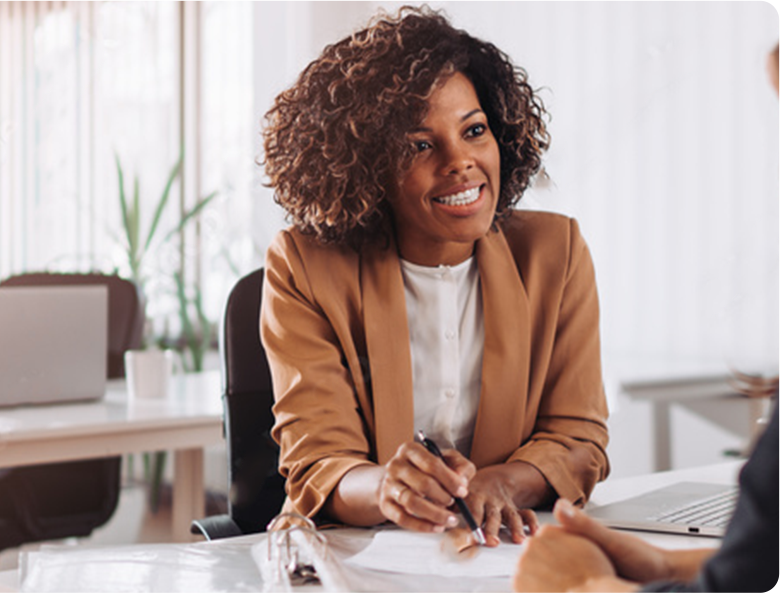 A woman with curly hair, dressed in a brown blazer and white shirt, sitting at a desk in an office, engaged in conversation.