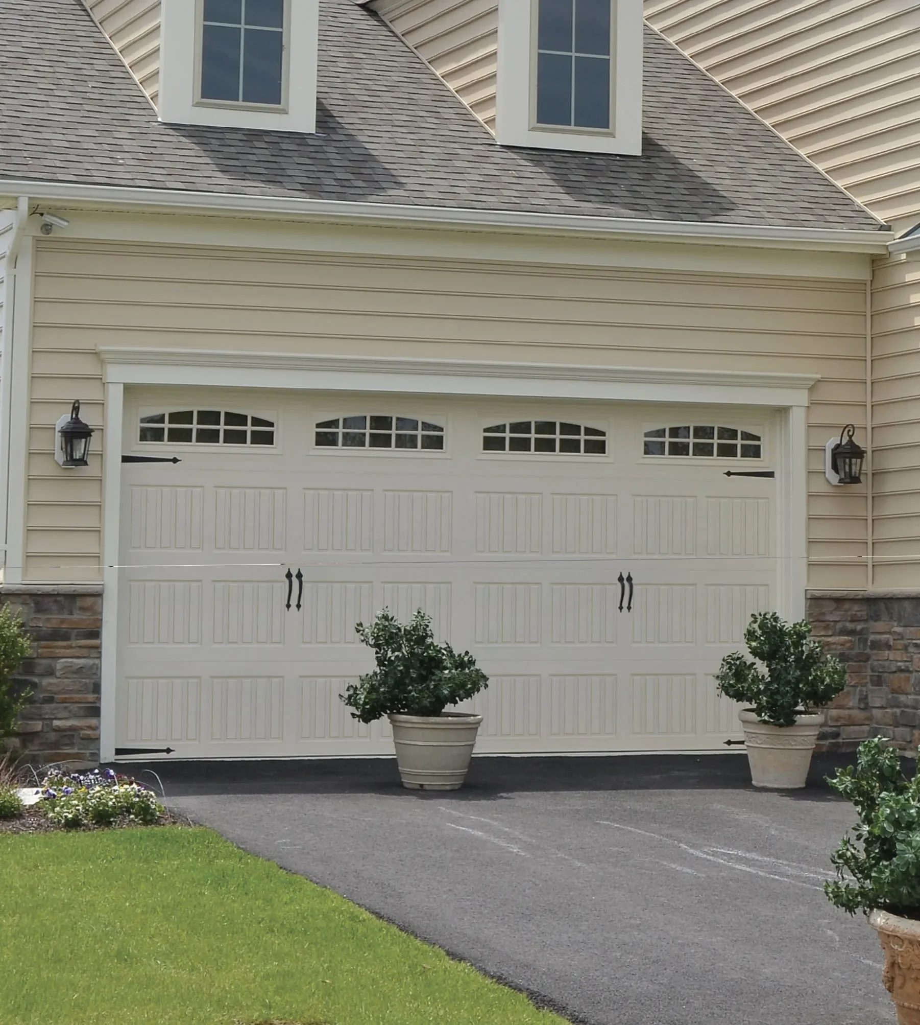 A white garage door with decorative windows at the top, flanked by black lantern-style lights, with two potted bushes in front, a black asphalt driveway, and a yellow house with stone accents.