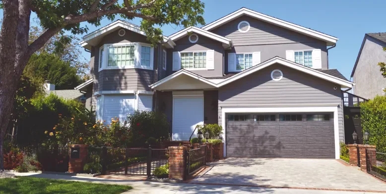 A two-story house with gray siding, a garage door, and a front yard with a brick fence and garden.